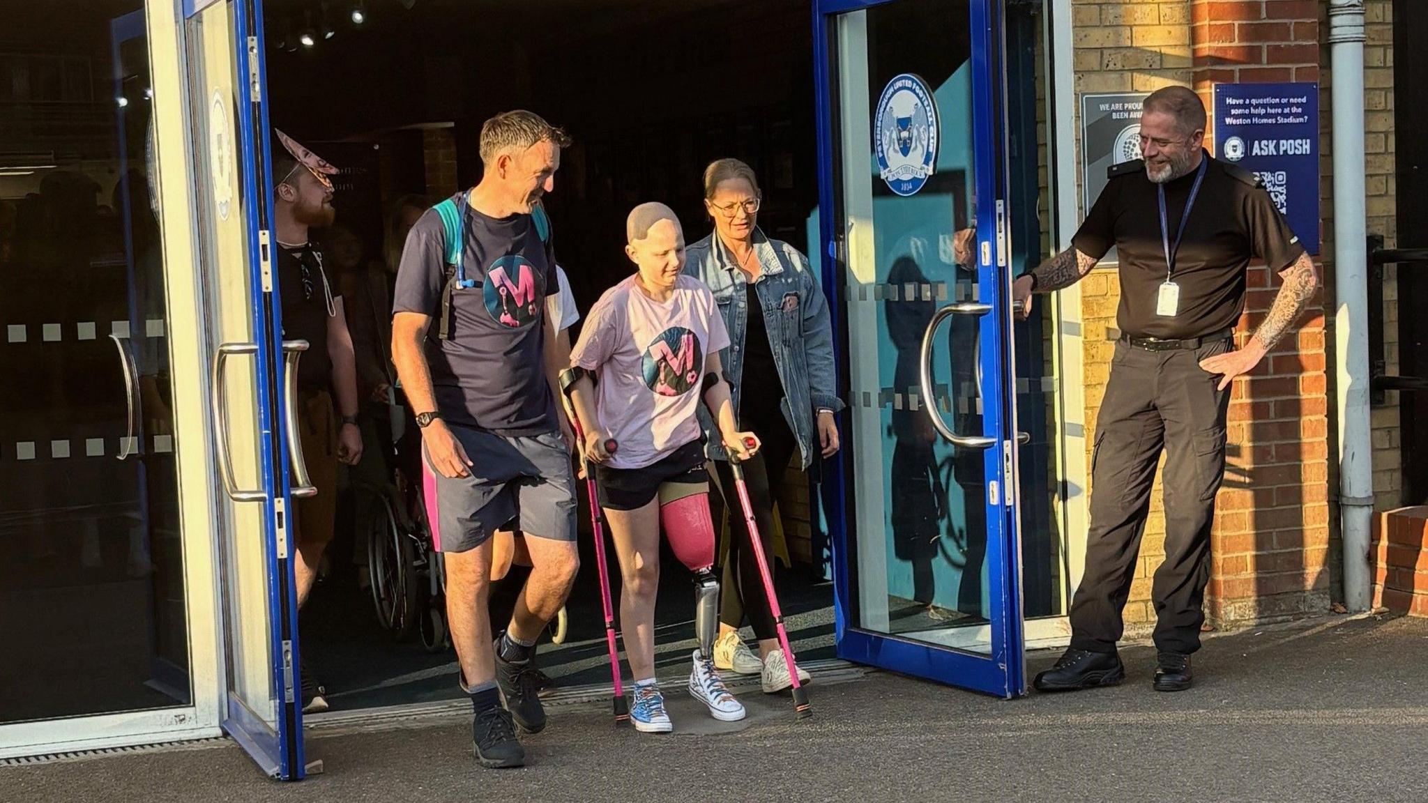 Millie on crutches walking out of the front door of a building. Her left left is a pink prosthetic thigh, rounded off at the knee. Her lower leg consists of a slim false section with a shoe. She is flanking by two people, with two others holding the double-doors open for them.