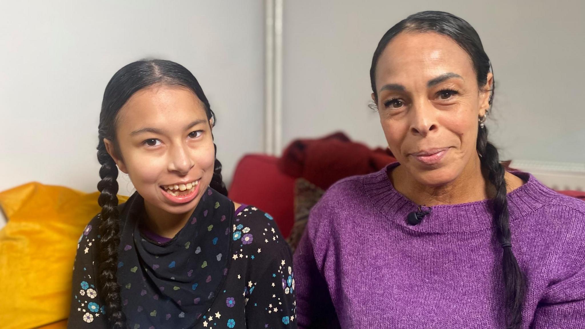 Neave and Natasha are sitting indoors on a sofa. Both have long, dark hair styled in braids/pigtails. Natasha, who is taller, is wearing a purple knitted top with a small microphone clipped near the neckline. Naeve is dressed in a black top patterned with colourful stars and flowers. They are both looking at the camera and smiling.