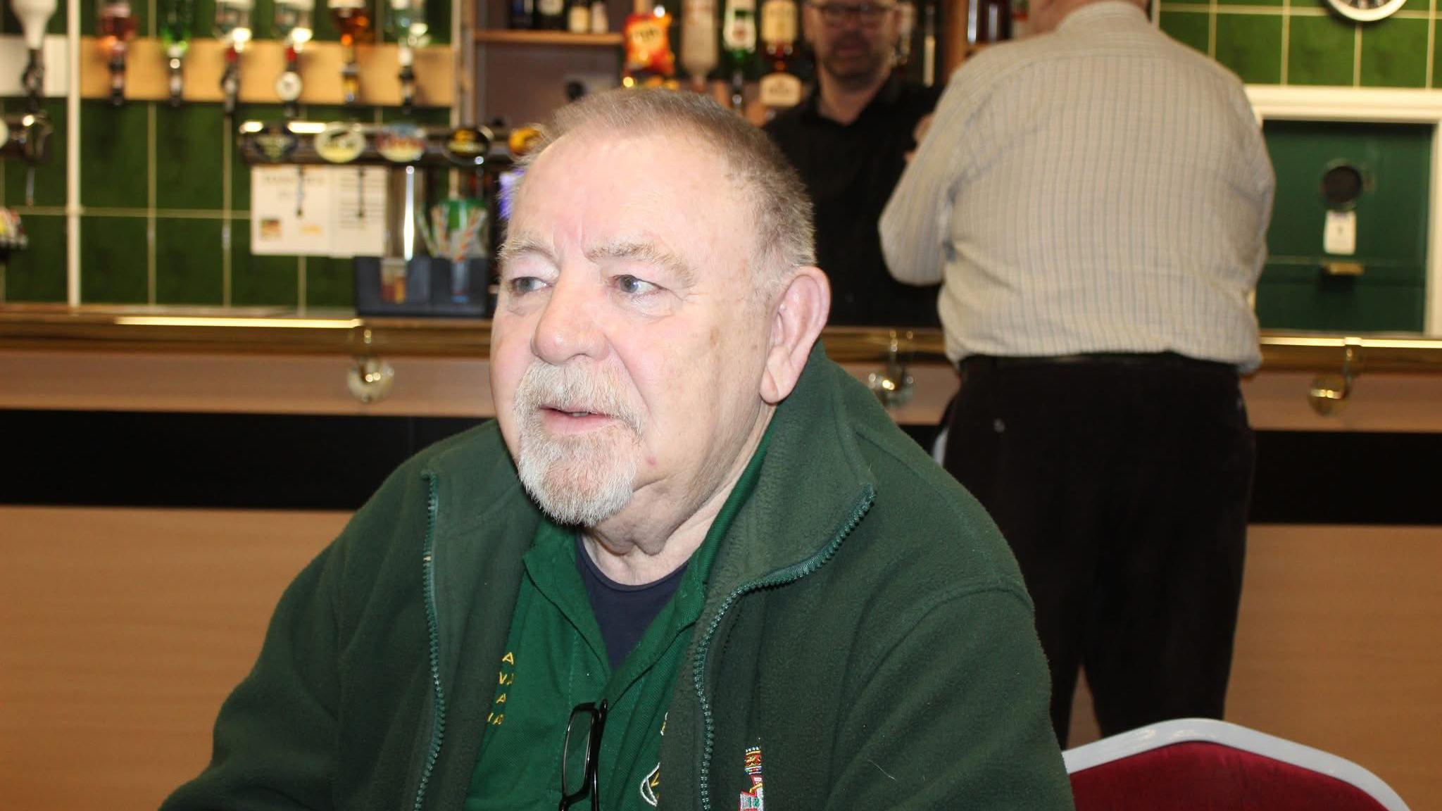 A portrait shot of Ray Bowen, who has received SABR treatment, sitting in a pub. He has a white beard and grey hair and is wearing a green fleece top and staring to the left of the picture.