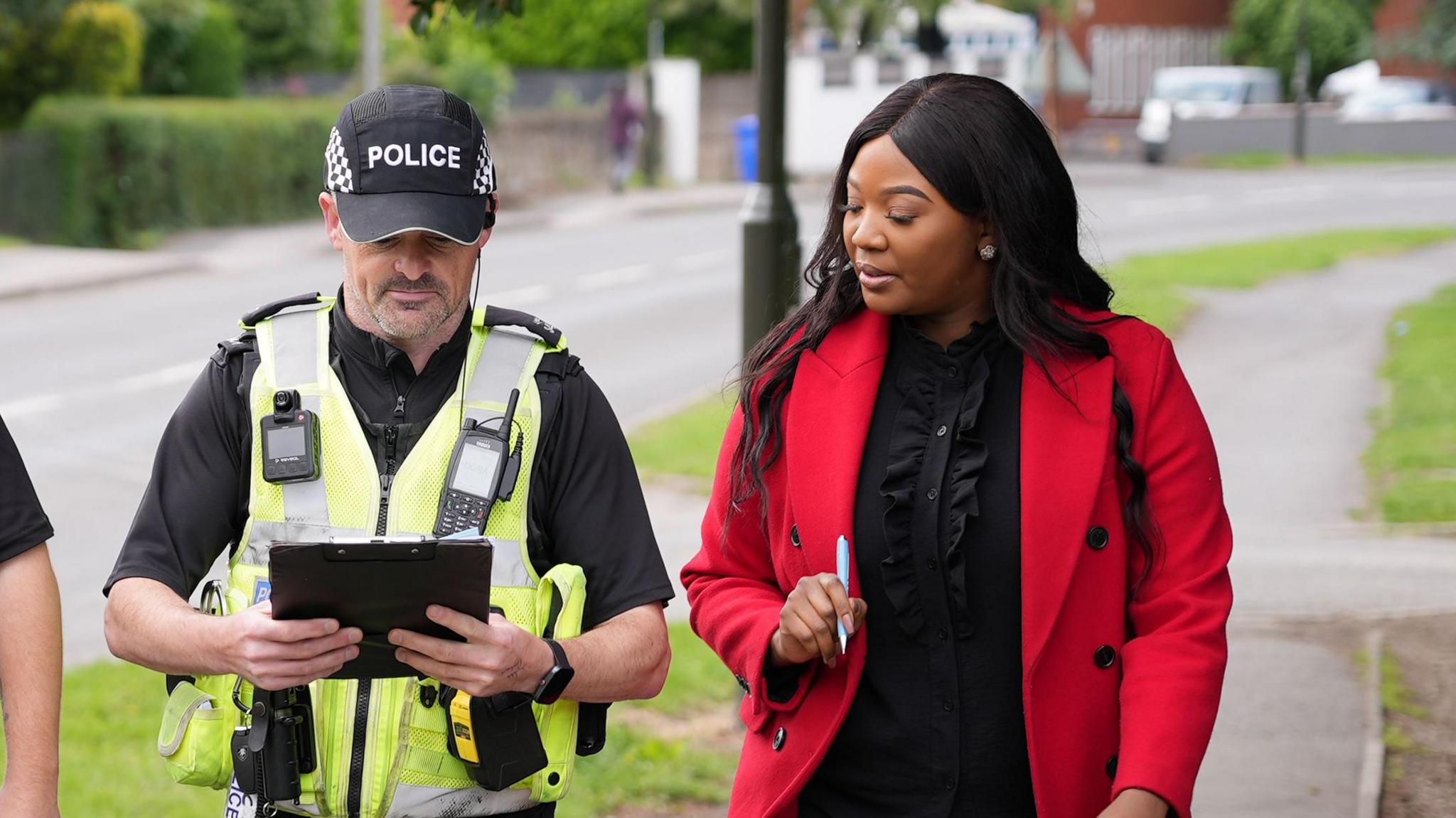Woman with black hair and red coat walks alongside police officer in neon vest carrying ipad in conversation