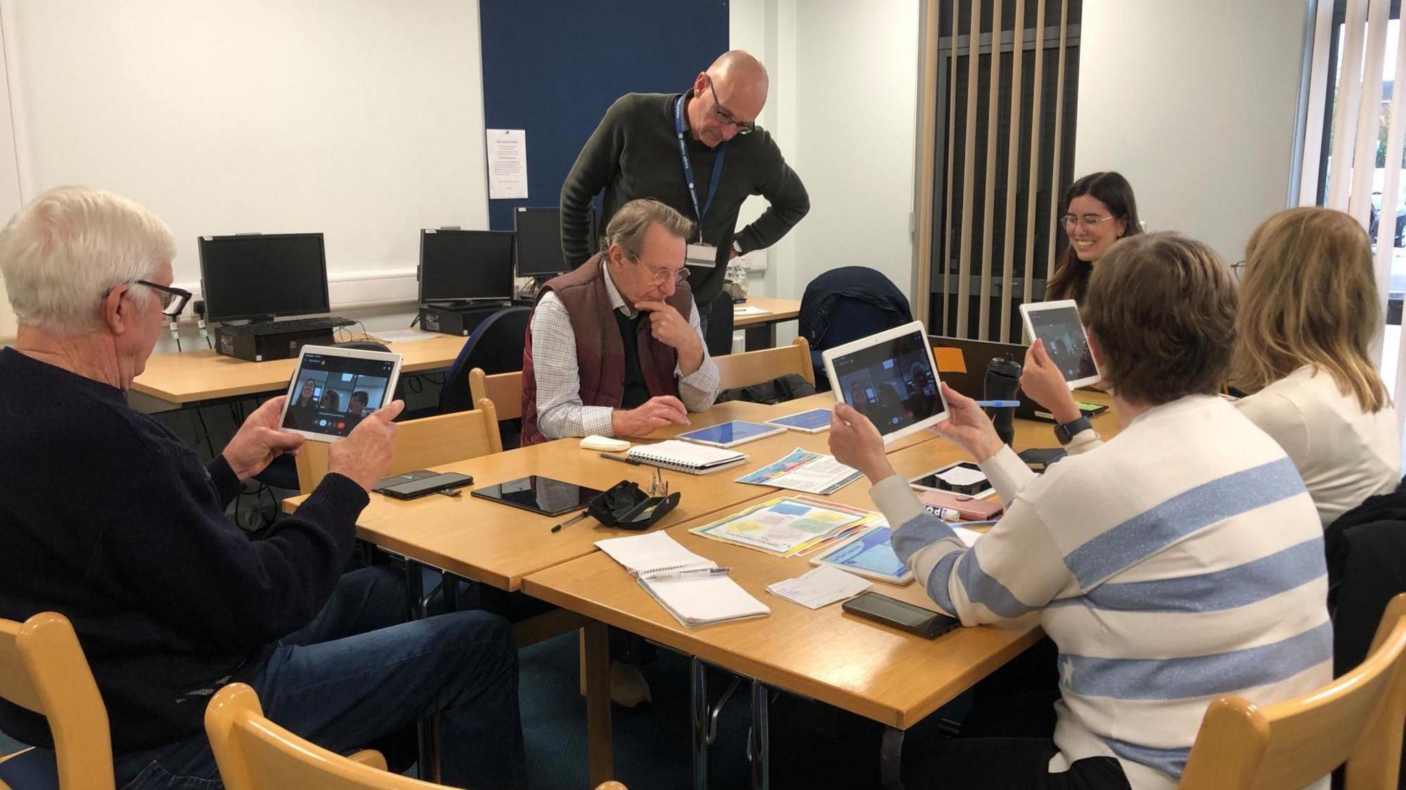 Five people are sat around a table and one man is standing near the table. Four of the people are looking at digital tablets on which there is a video call. In the background there is a row of three desktop computers.