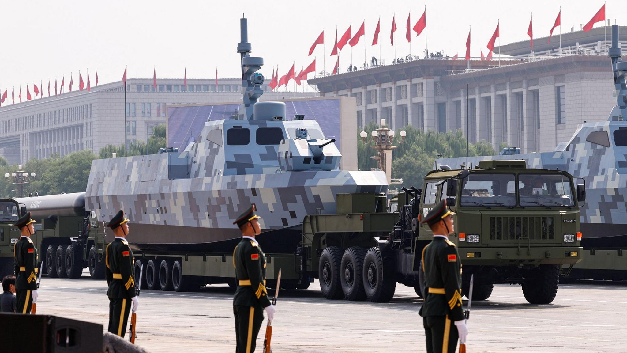 A camouflaged patrol boat on the back of a transporter at a military parade with Chinese soldiers standing to attention in the foreground
