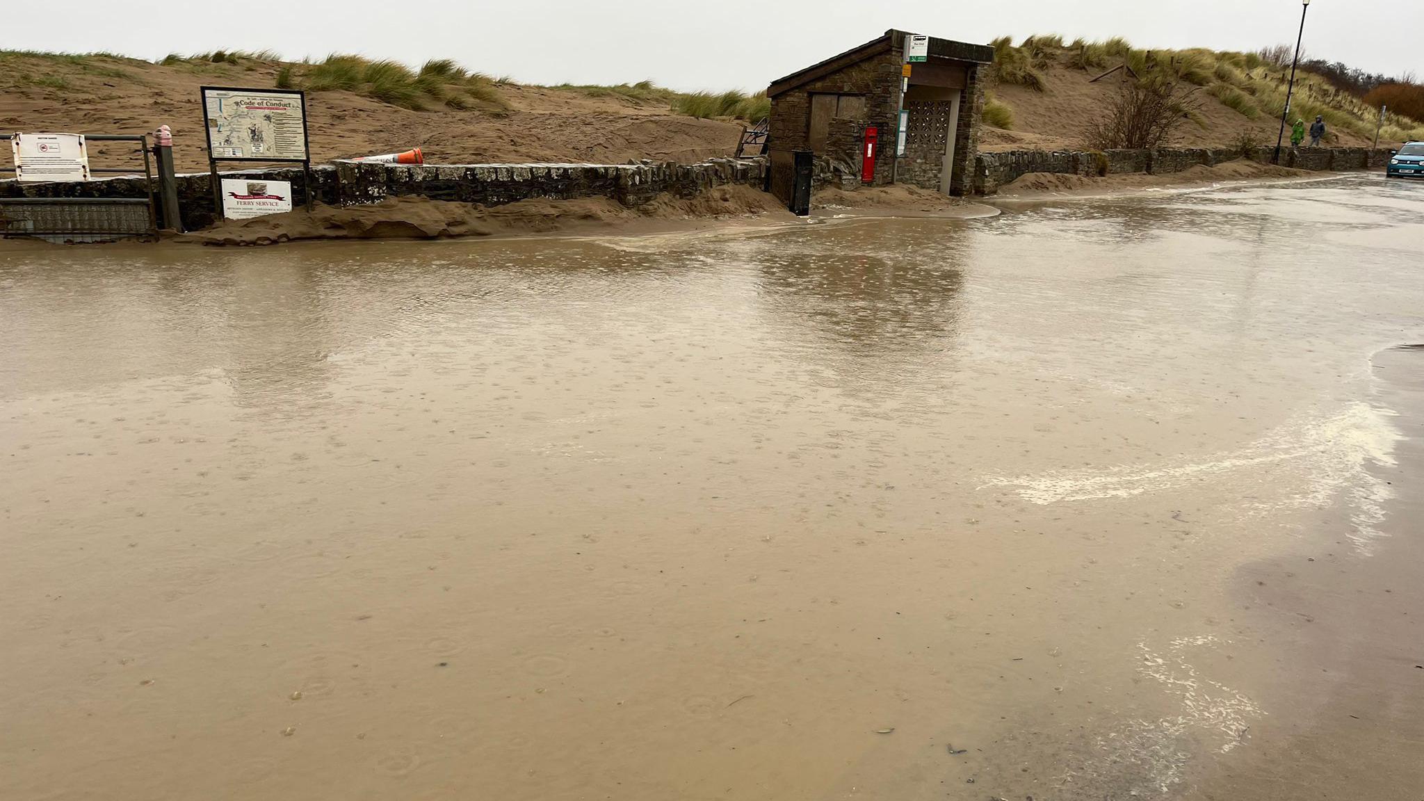 Flooding on Marine Parade in Instow on a rainy day. Sand dunes are in the background along with a bus stop shelter and red post box.