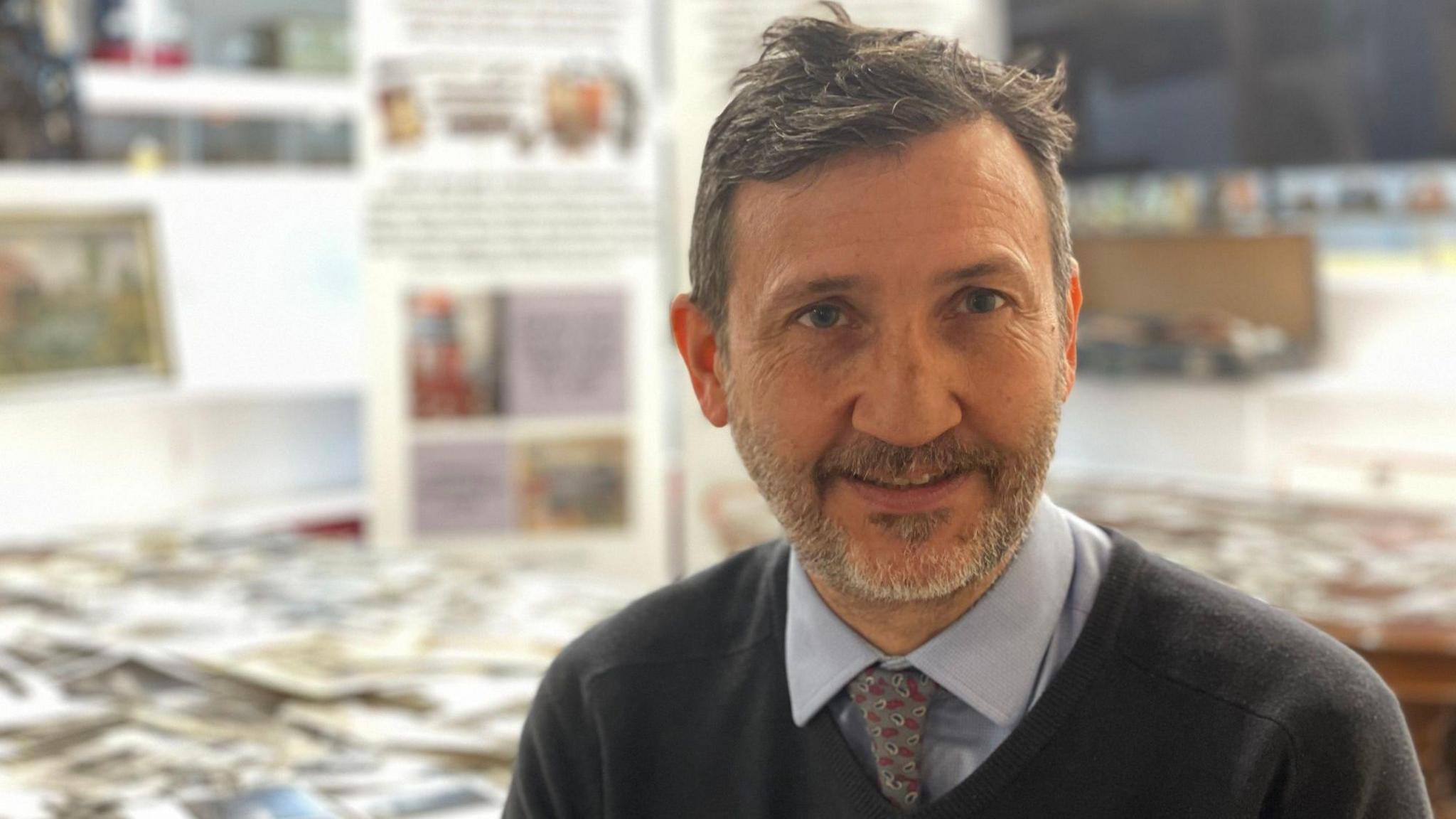 Darryl Kirk in an office with hundreds of photos spread across a table. He has short dark hair and a short beard. He wears a blue shirt, grey and red tie, and a dark grey jumper.