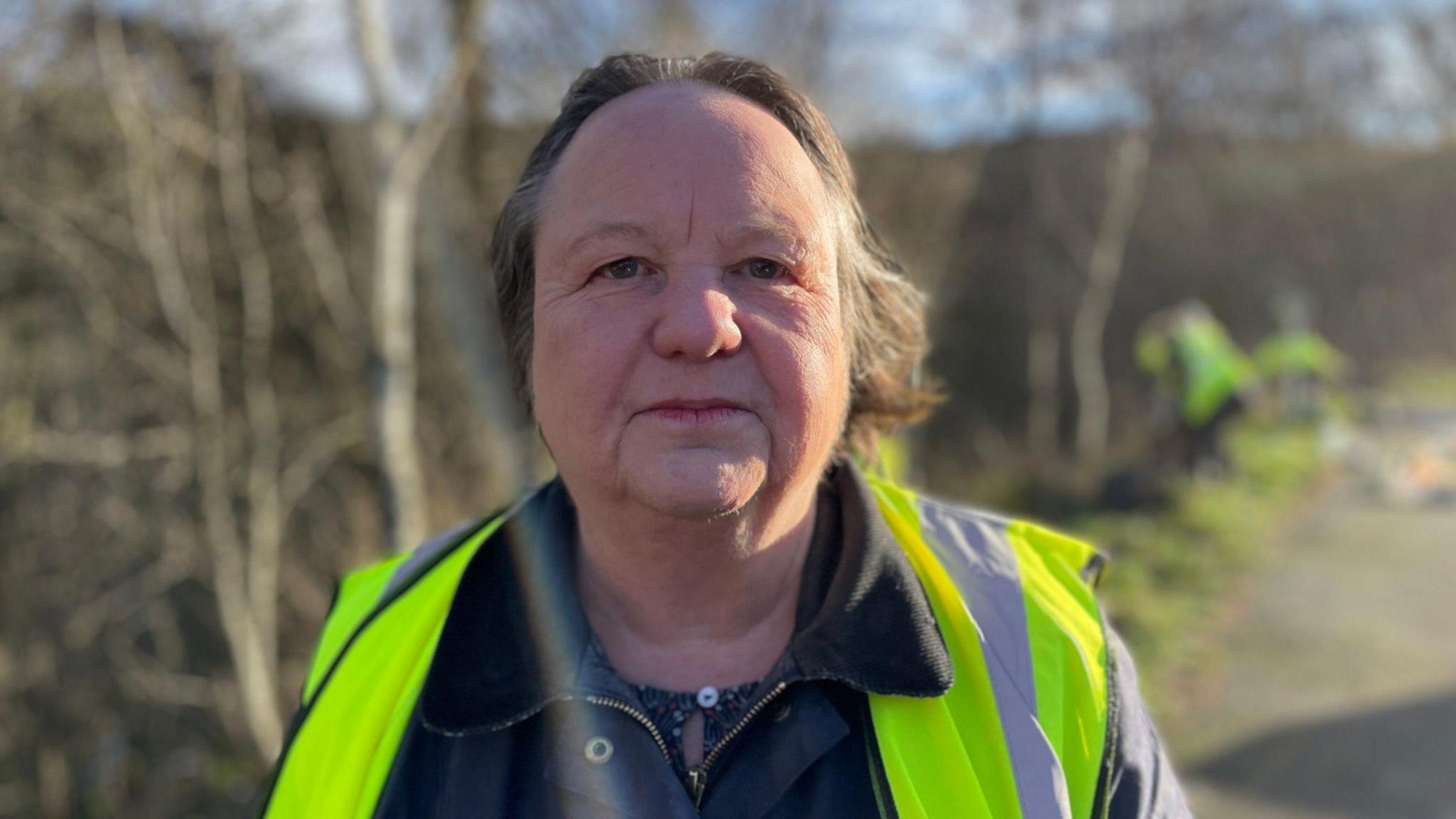 A woman with a high-vis jacket and wax coat looks at the camera, while litter pickers are collecting rubbish from the undergrowth in the background