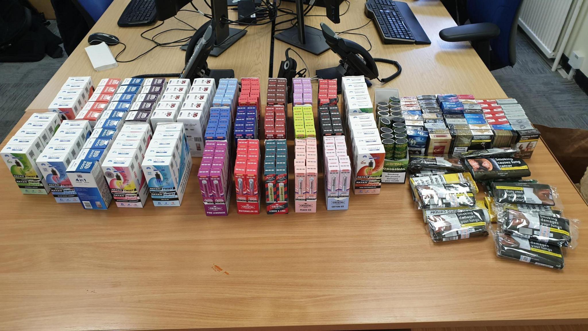 Hundreds of suspected illegal vapes and counterfeit cigarettes lined up on a wooden desk. The boxed vapes come in a variety of different colours. Computer, a keyboard and a phone can be seen at the back of the desk inside an office.