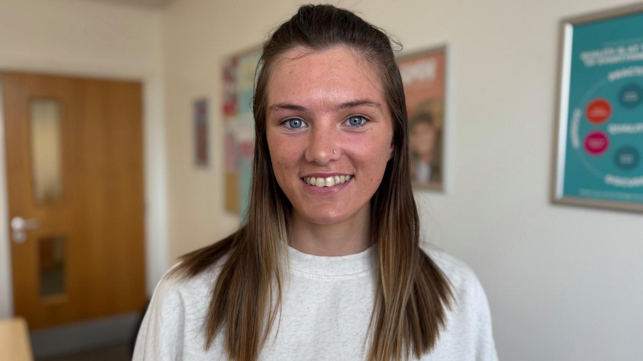 Close-up portrait of Neala with shoulder-length brown hair wearing a light-coloured sweatshirt, standing indoors. A wooden door and framed posters are visible in the softly blurred background.