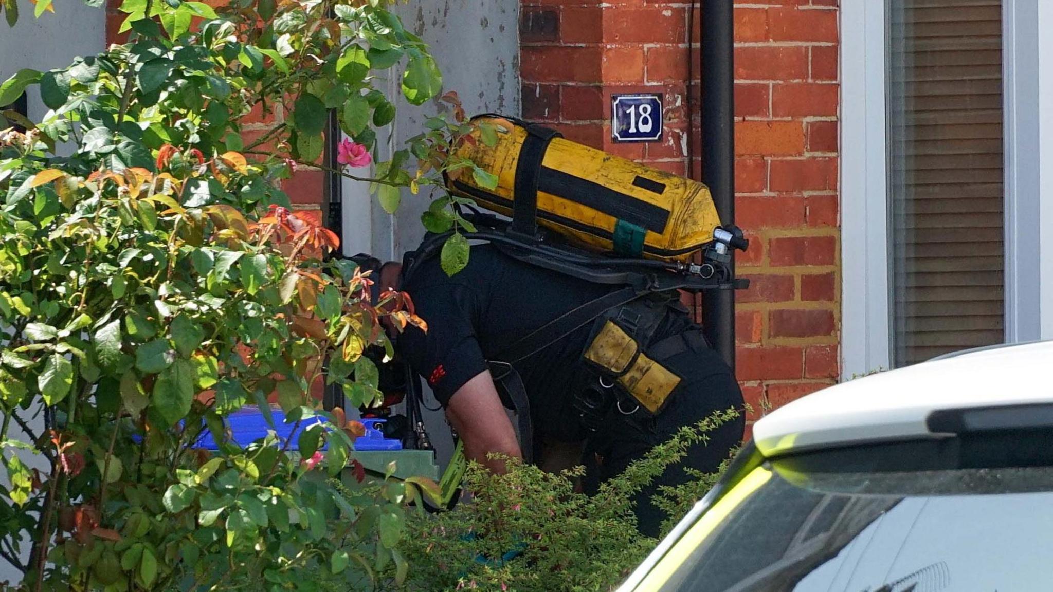 A close-up of a man dressed in black wearing an unknown yellow tank on his back and a contraption which looks like a gas mask over his face bends over on the doorstep of a brick build terraced house.