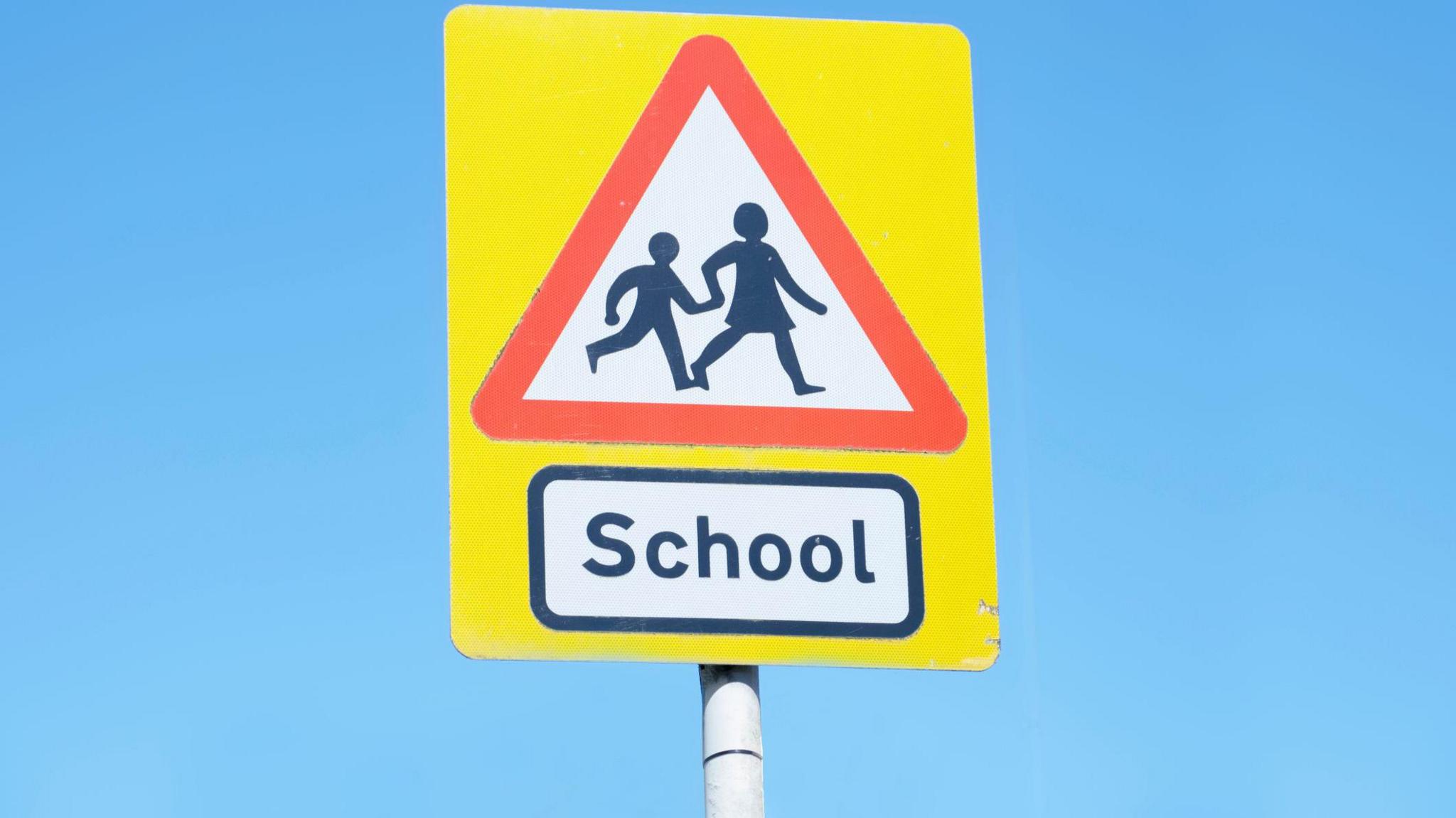 A yellow road sign which has a red triangle on it and a picture of two people holding hands and crossing the road. Underneath the triangle there is a rectangle with the word school written in it in black letters.