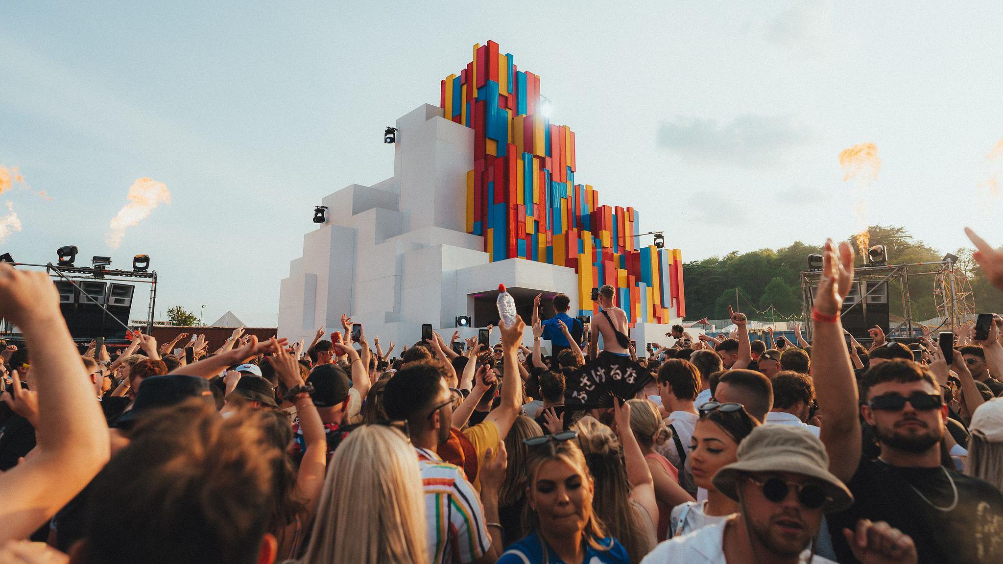 A large crowd of people gathered in front of a DJ booth at Love Saves the Day festival. The stage is a large geometric patterned structure which is white on one side and multicoloured on the other. The crowd are waving their arms in the air and the sky is a very bright white.