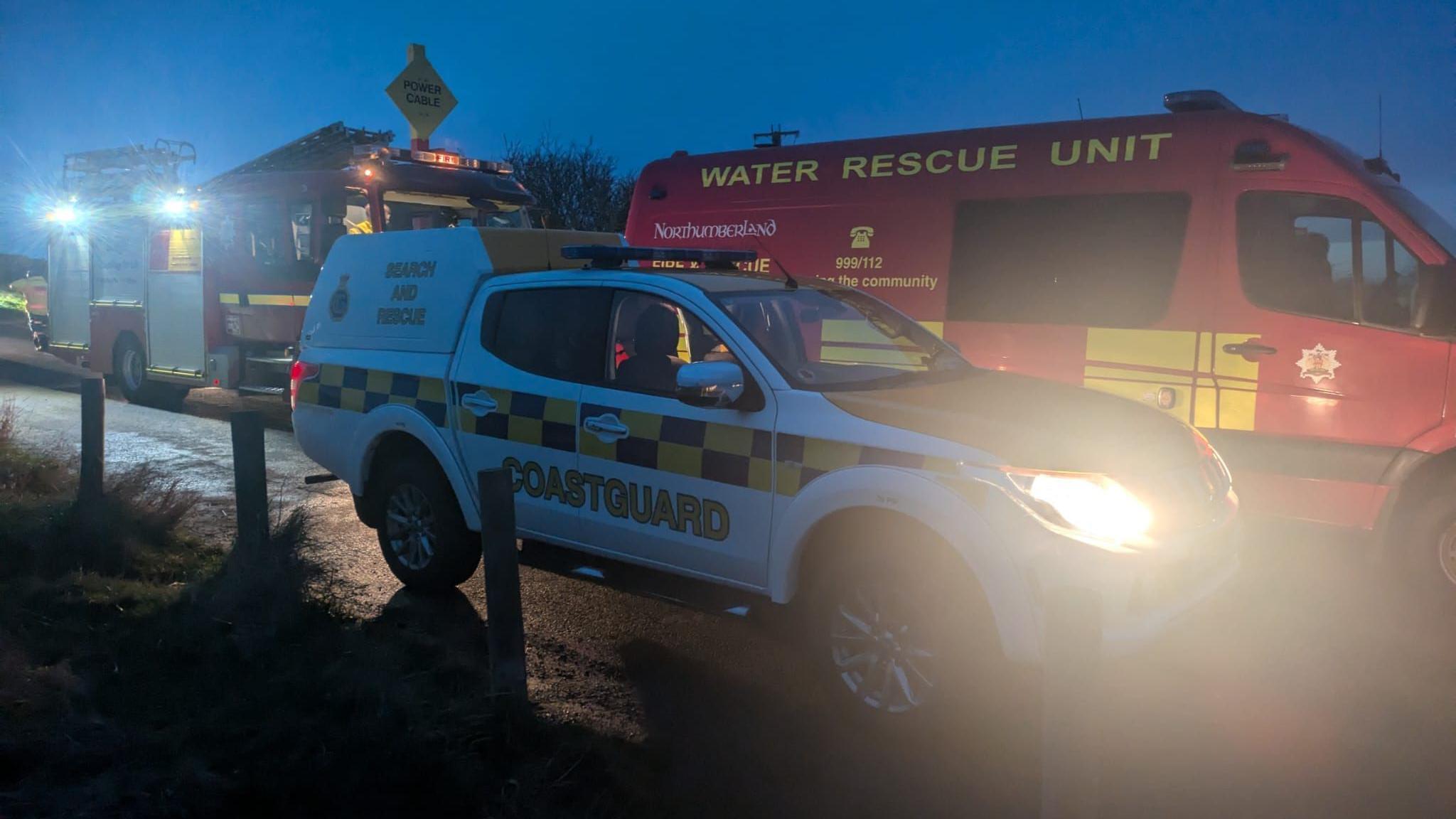 Coastguard and fire service vehicles parked on a narrow road in the dim light. 