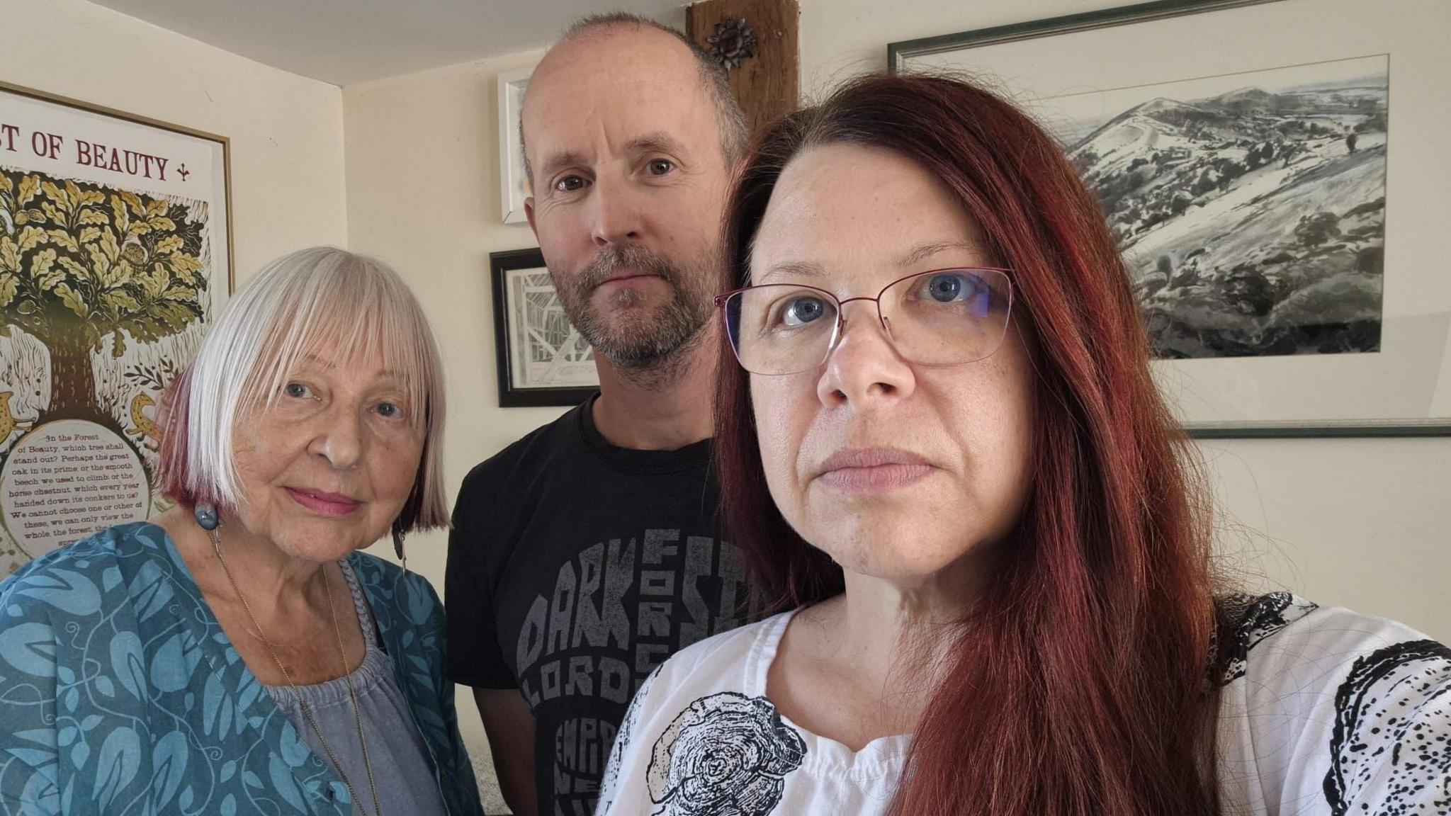 A woman with long brown hair and glasses takes a selfie of herself with two others. She is wearing a white top with black flowers on it. Next to hear is a man with a brown beard and short greying hair wearing a black t shirt with grey print on it. To his right is an older woman with bobbed white hair and blue floral tunic. They are in a living room with cream walls and various framed prints.