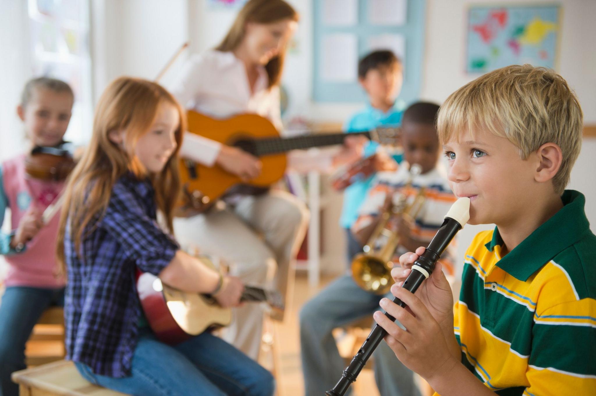 A young boy plays the recorder in a school setting with other children playing guitars, trumpets and violins behind him.