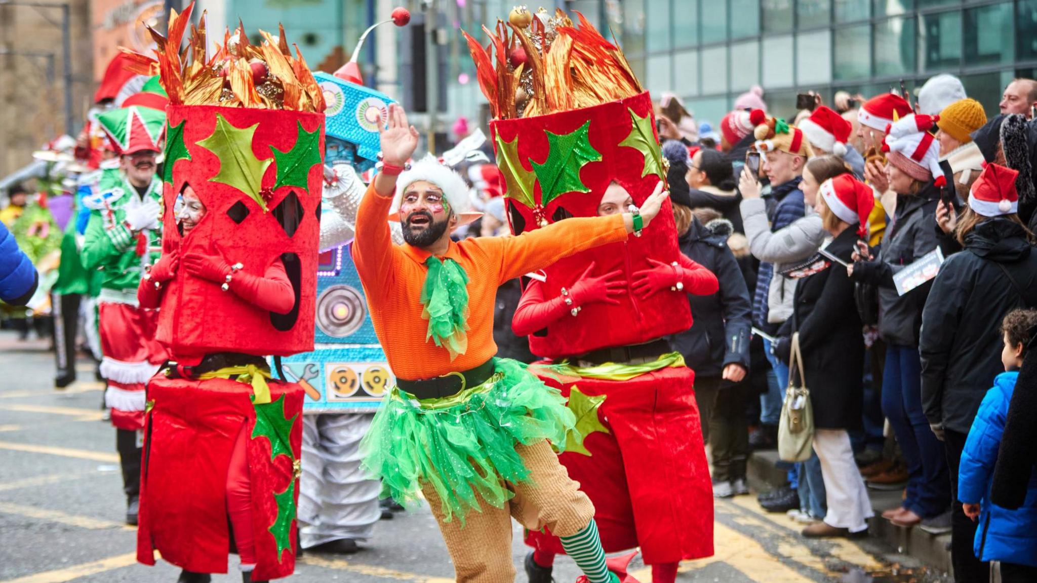 A man in an orange and green elf outfit waves and dances in front of people dressed in large red cracker outfits as they parade past a line of spectators in santa hats