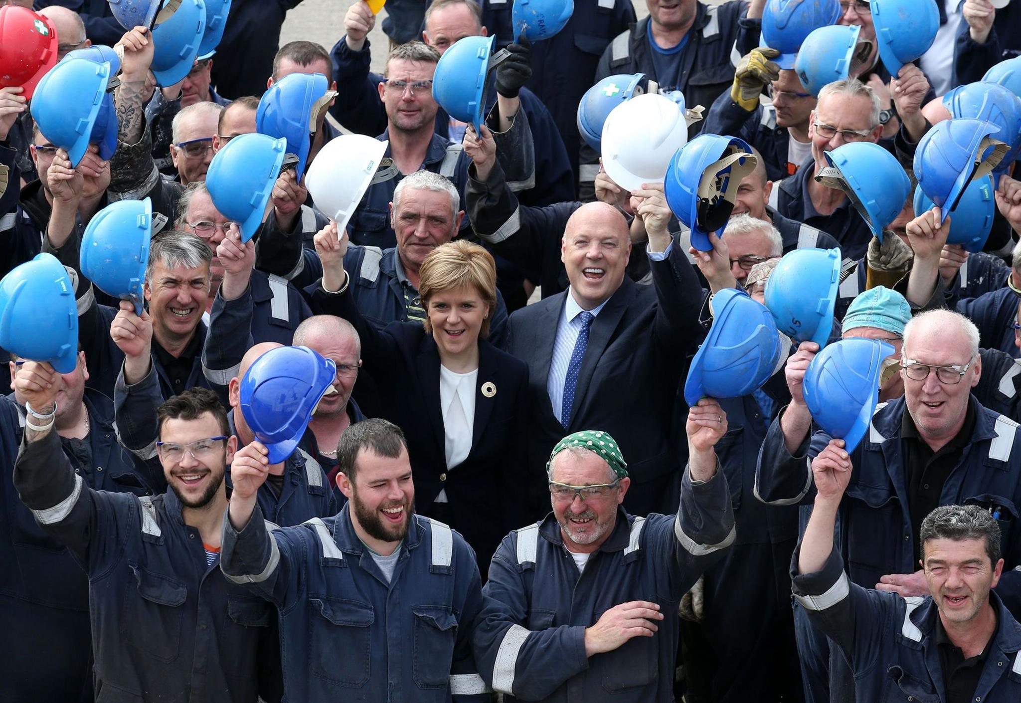 Nicola Sturgeon and Jim McColl, a bald man in a dark suit surrounded by shipyard workers raising their blue helmets in celebration as Ferguson's shipyard is announced as preferred bidder for the ferry contracts