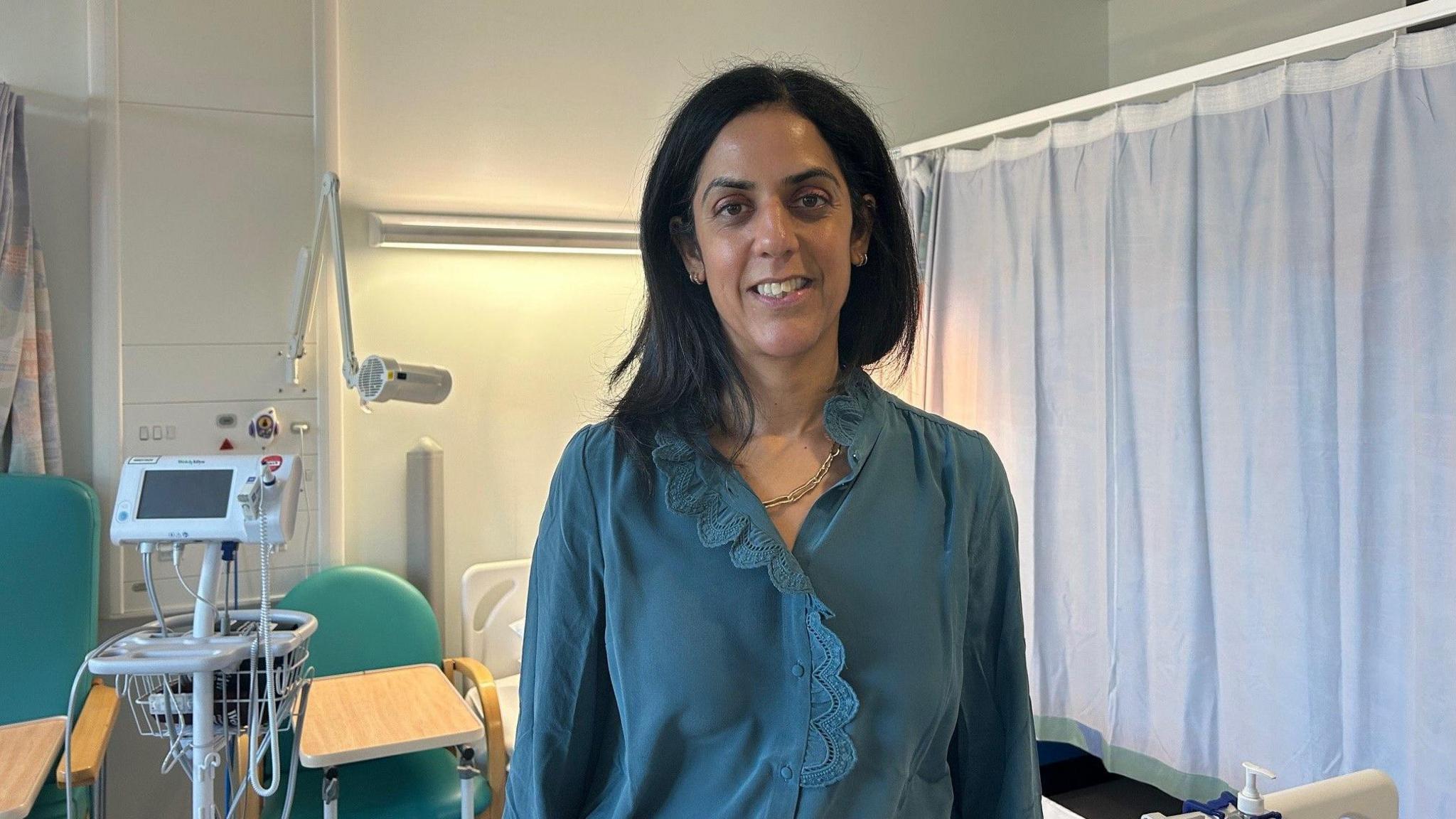 A woman in a blue blouse smiling in a hospital room.