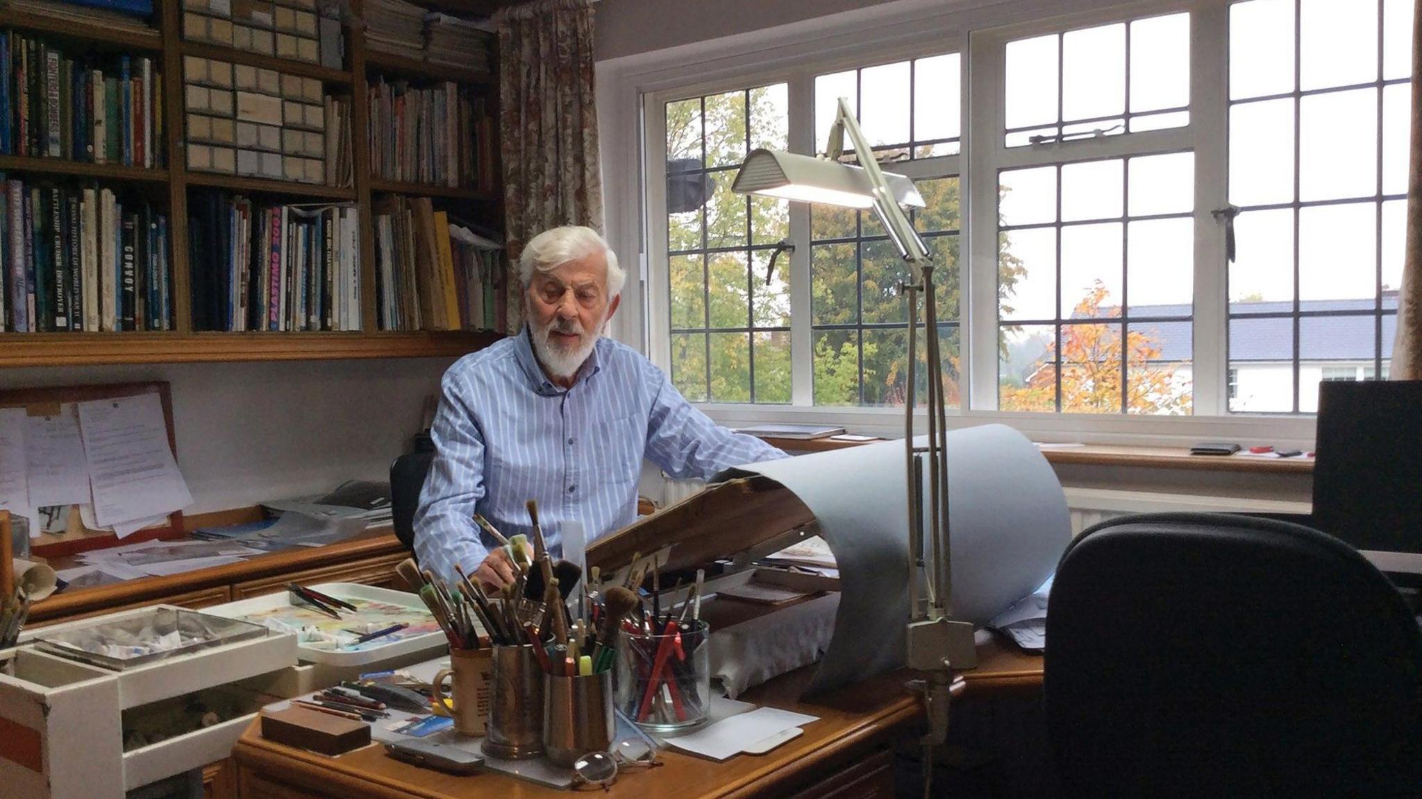 A grey-haired man, with a grey beard, wearing a blue-and-white-striped shirt and sitting at a desk in front of an easel on a desk in a study. Trees with green and golden leaves are visible out of a window to the man's left. Books line the wall behind him. Jars filled with paint brushes can be seen on the desk.