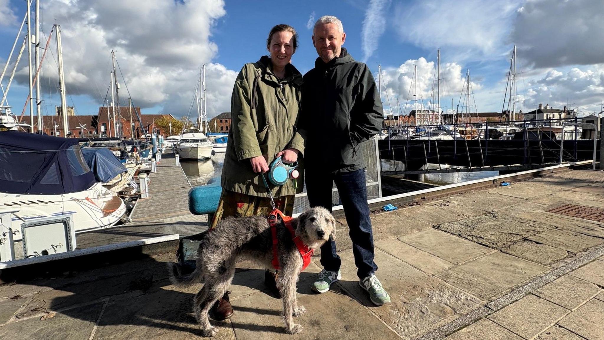 A middle-aged couple are pictured with their grey lurcher dog, with boats moored on a marina behind them. The woman is wearing a long, olive green jacket. Her husband is wearing a black jacket and navy jeans.