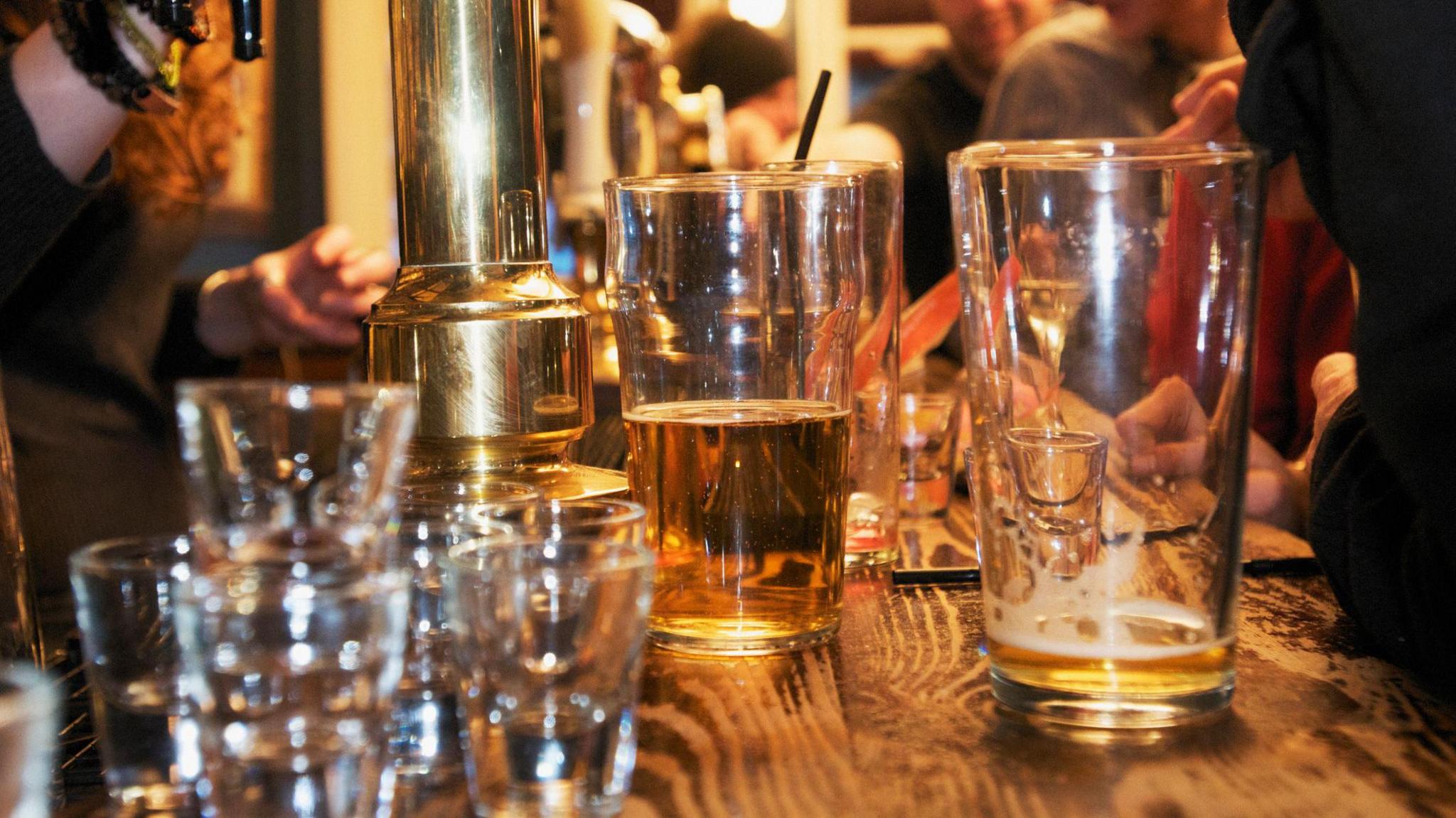 A stock photo showing a wooden bar top with a beer bump. The bar is covered in glasses of different sizes, some partly full of drink and some are empty