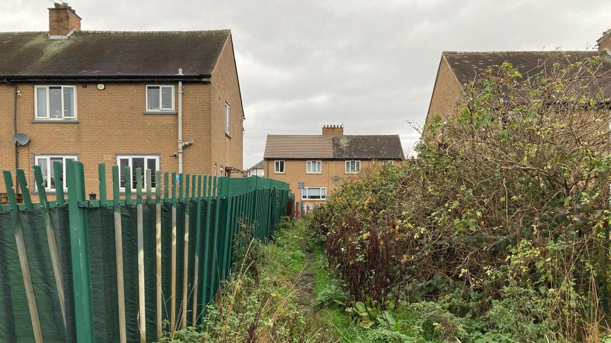 A ginnel flanked by a green metal fence going in between two semi-detatched brown brick houses