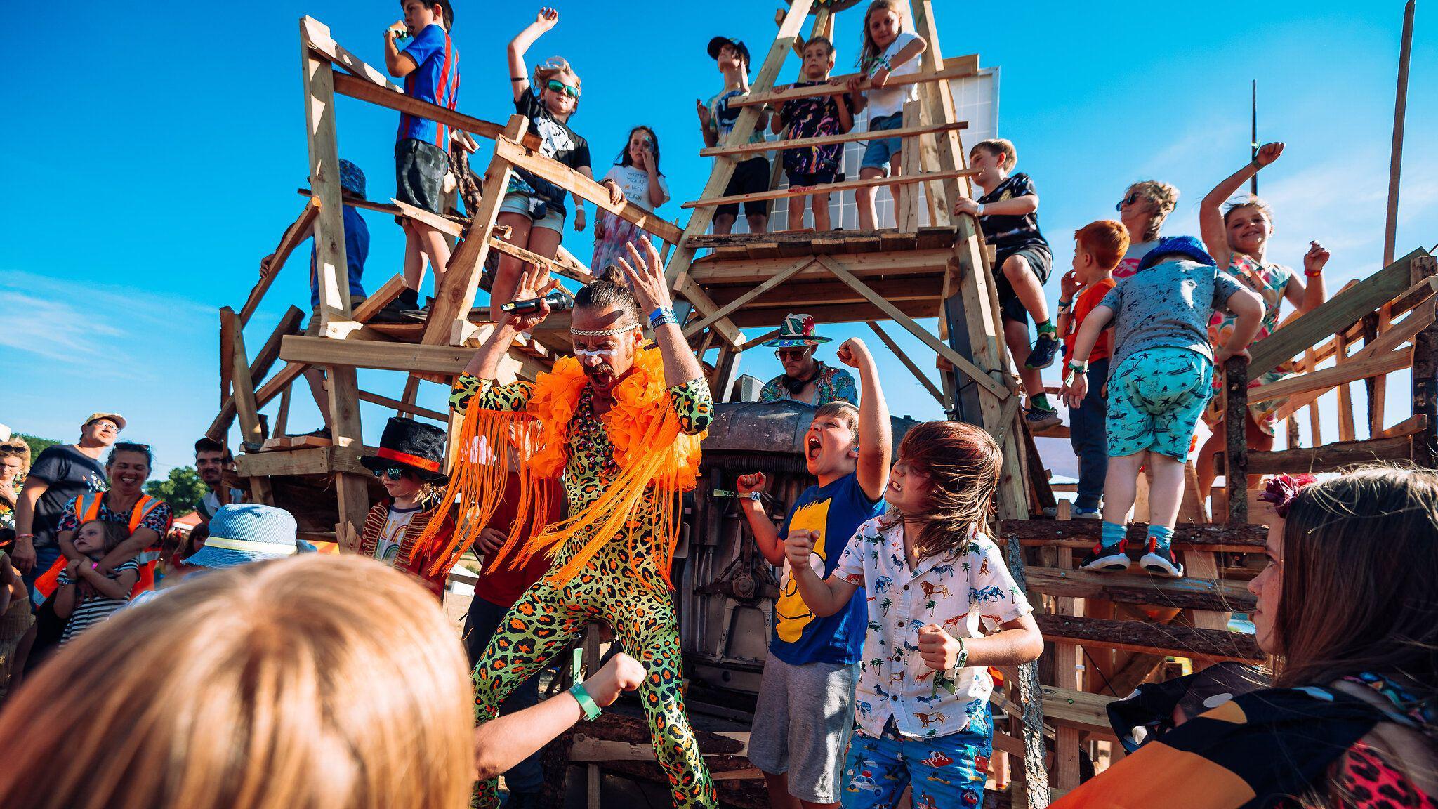 A man in a green and orange leopard print catsuit with an orange shawl and children dancing and singing next to him. There is a wooden climbing stand behind them with children climbing on it and a DJ at a booth within the wooden construction. 
There is a crowd of people to the sides and behind the camera and the sky is bright blue.