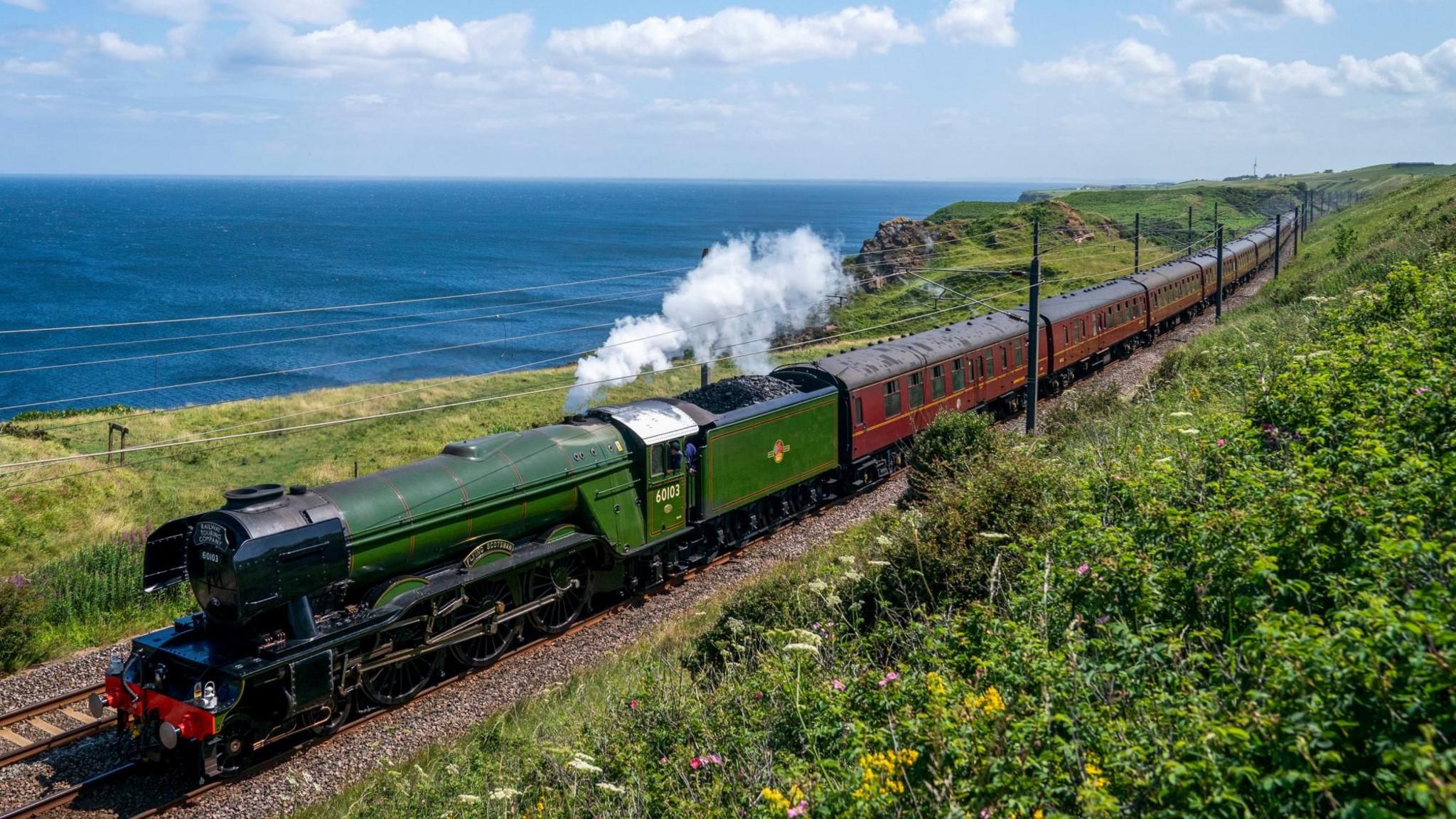 A green steam locomotive passes through verdant countryside with the sea in the background and a blue sky with white fluffy clouds above. Steam is rising from an outlet of the locomotive
