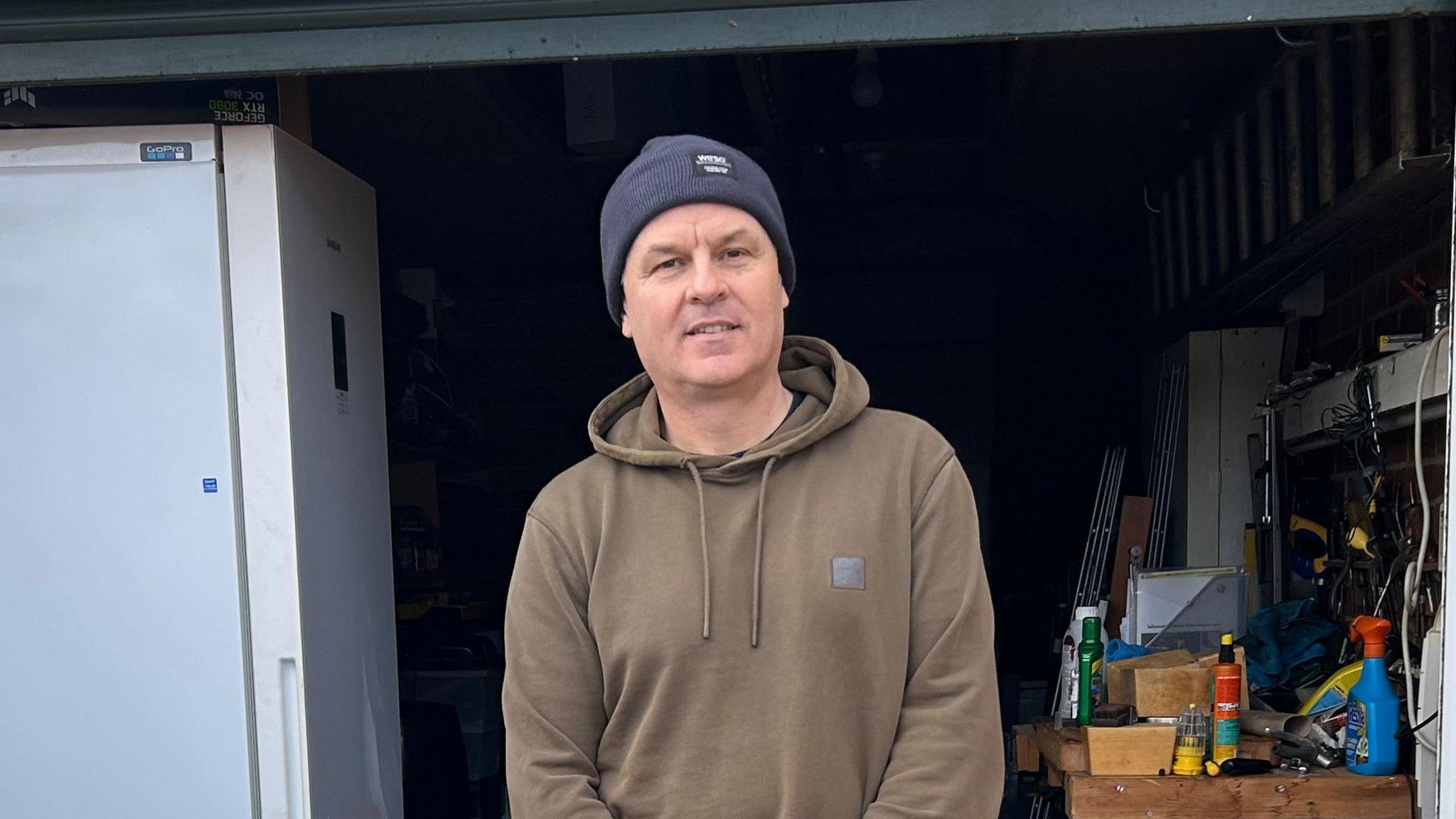 A man smiles at the camera standing in front of an open garage. He is wearing a navy beanie hat with a brown hoodie on.