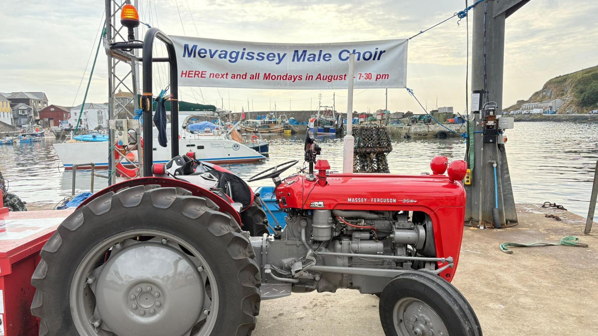 A red Massey Ferguson 35x tractor in front of a sign that says "Mevagissey Male Choir". The harbour is behind. There are boats in the harbour. The sea is clear. The sly is light blue with clouds.