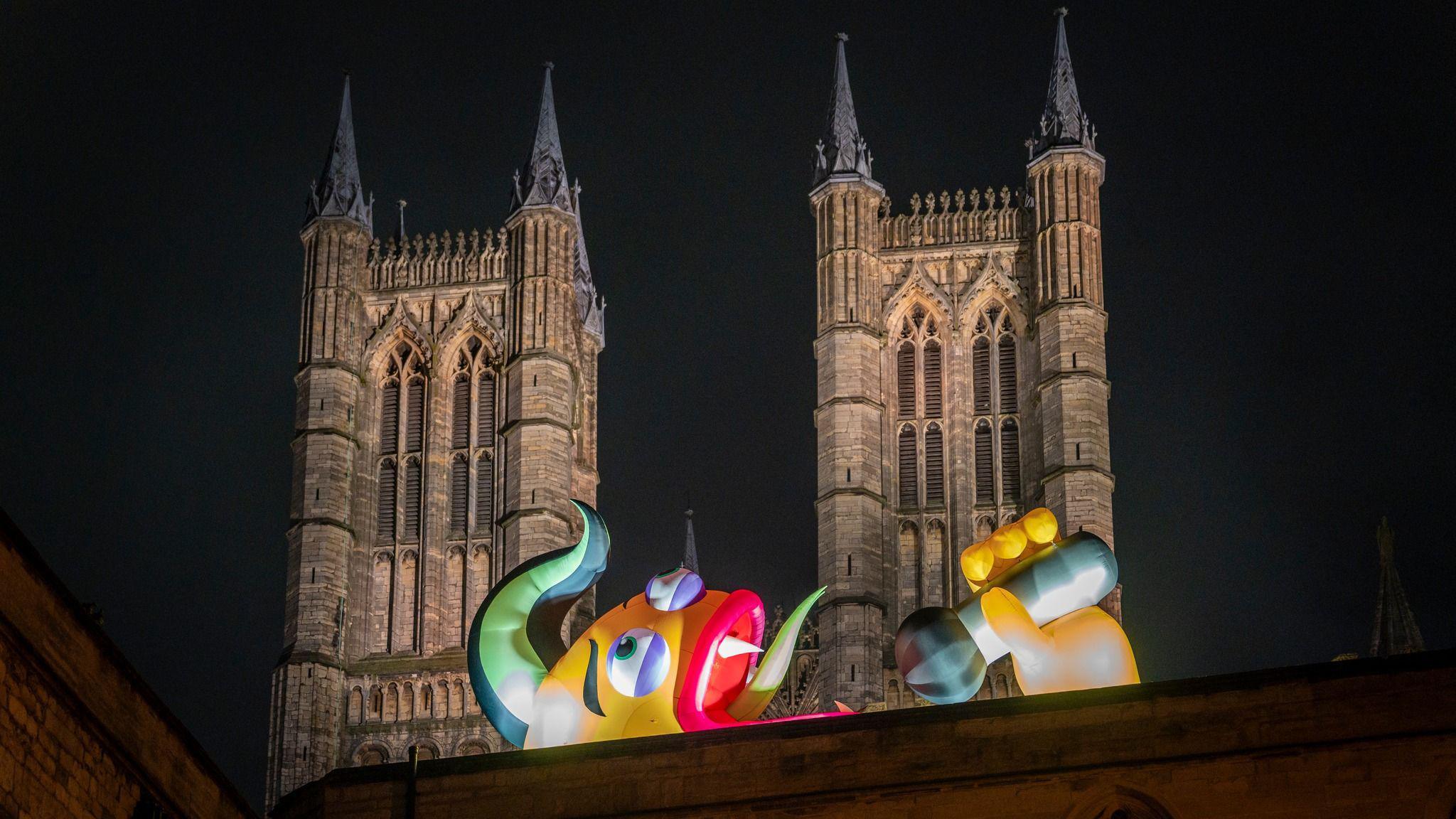 An illuminated yellow, green, purple and red inflatable monster on top of a wall and in front of two towers of a cathedral. It has a green tuft of hair and is lying on its side with its mouth open with a green snaky tongue and is holding a microphone in its yellow hand, mimicking being in mid-song.