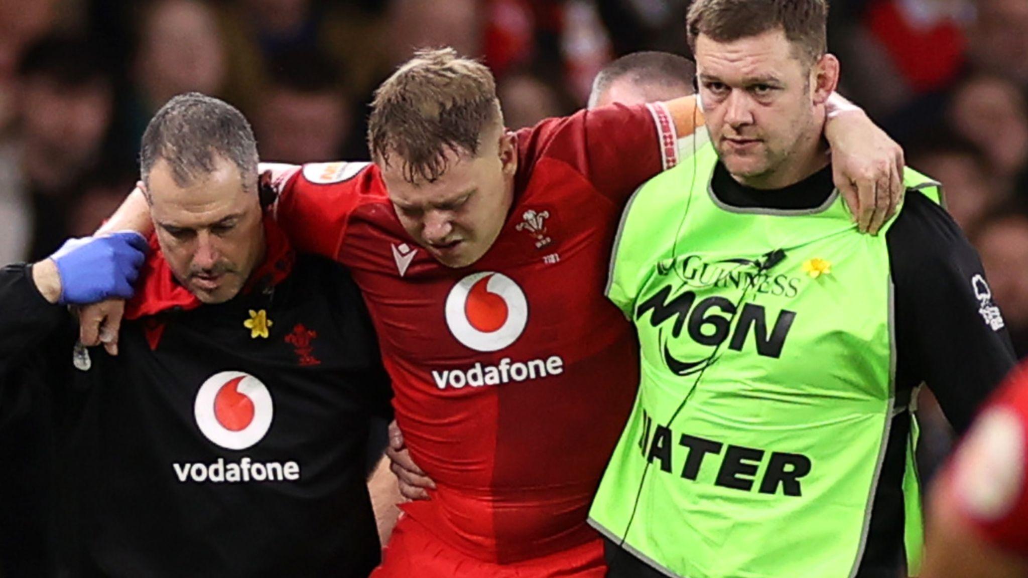Sam Costelow with arms around medics looks in pain as he is helped from the field