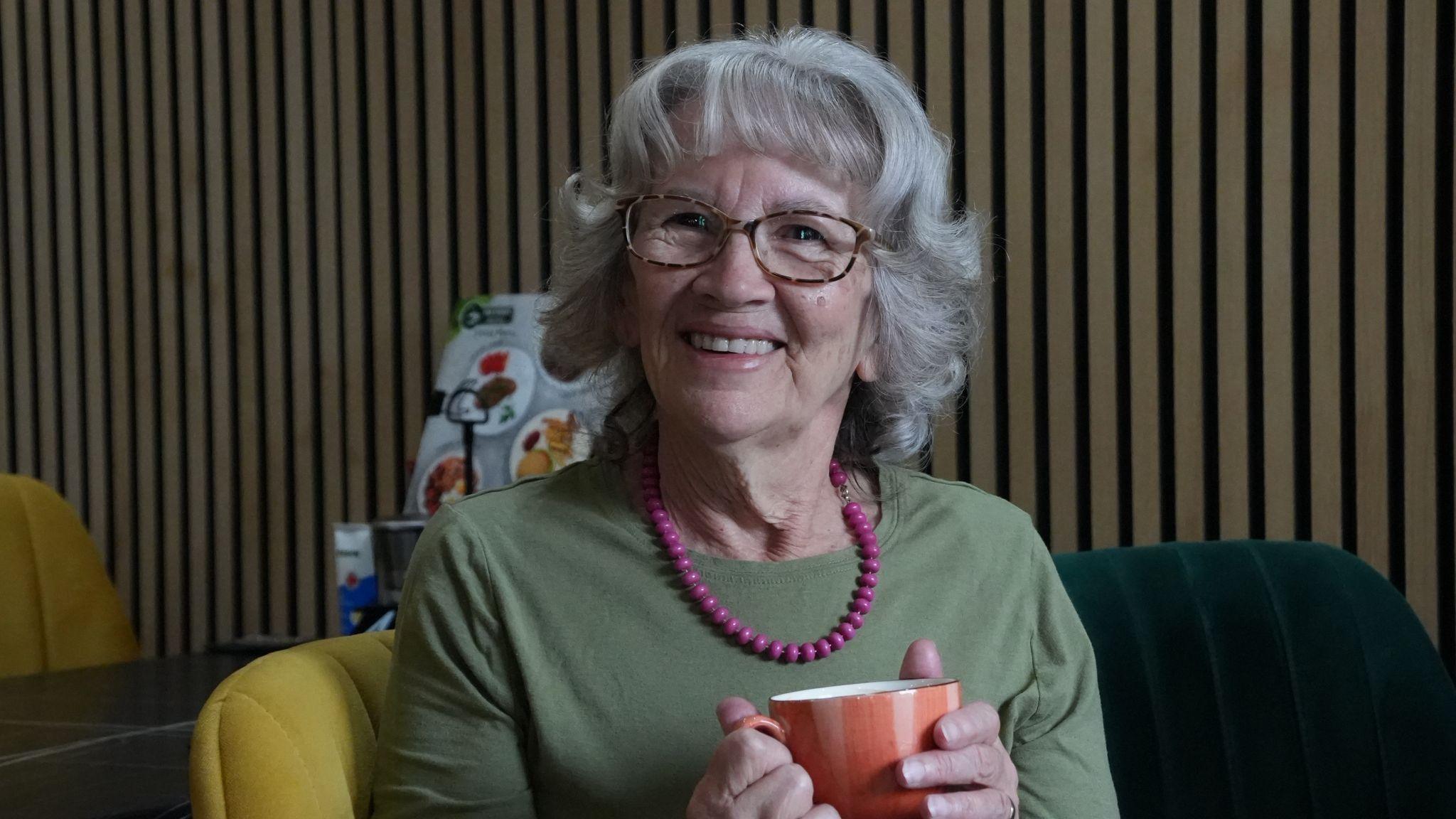 Margaret Davy is sitting on a chair in a restaurant with a coffee mug in her hands. She has wavy grey hair and is wearing spectacles, a green top and has a pink beaded necklace on
