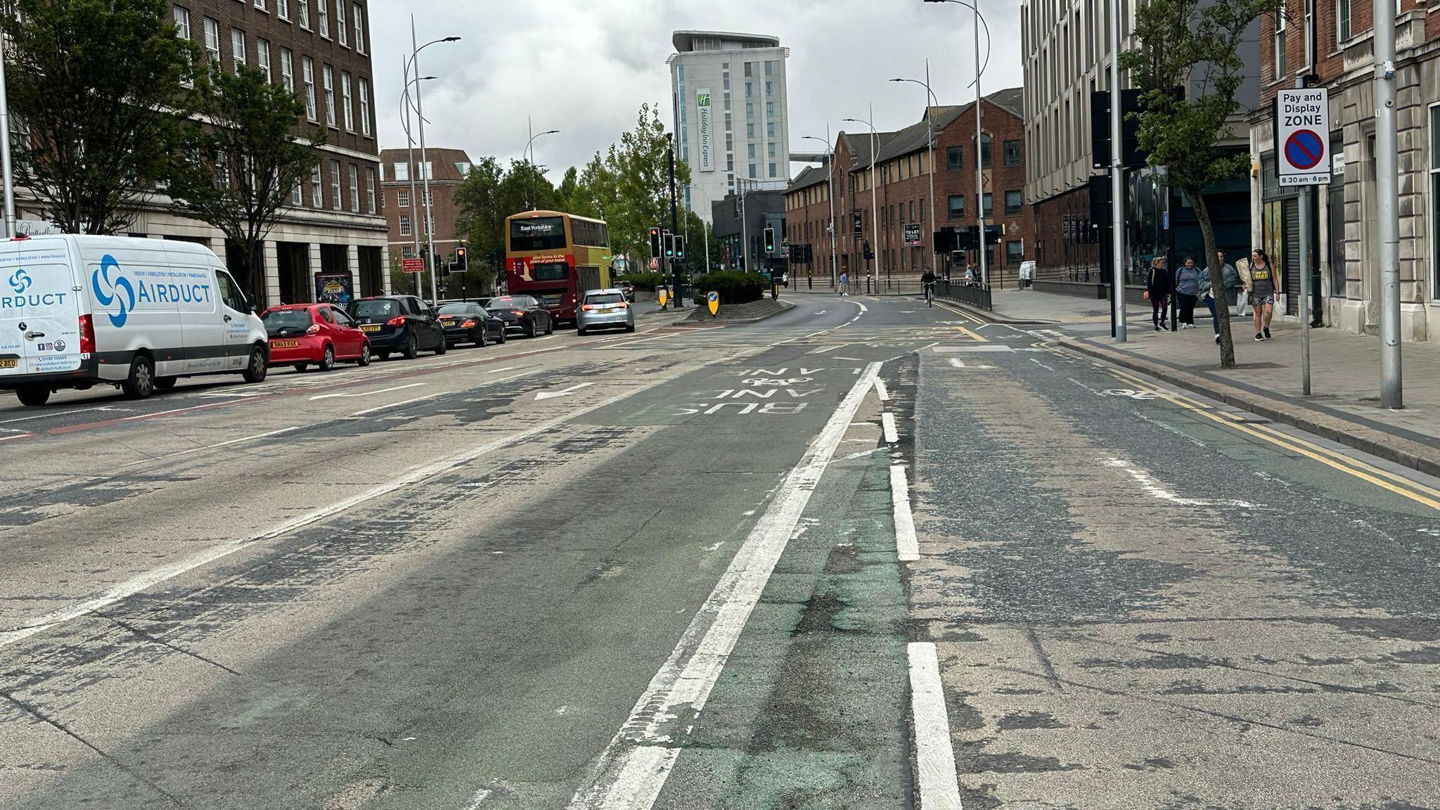 A city centre road with a bus lane on the right and several cars, a white van and a bus waiting at traffic lights on the left. Several brown and beige coloured buildings are either side of the road with a few people walking on the pavement.
