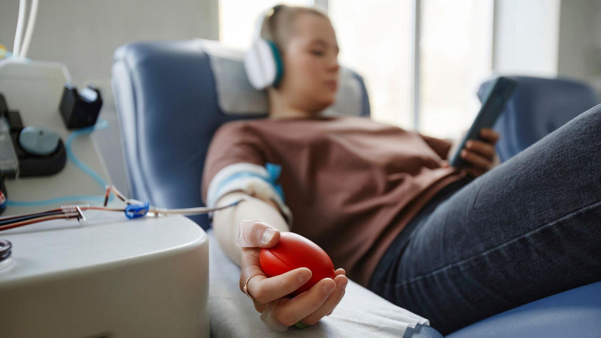 A stock photo of a woman sat in a blood donation chair. She's wearing dark blue jeans and a brown jumper, and has a pair of headphones in and a mobile phone in her left hand. Just her right hand is in focus - where she's squeezing a stress ball to keep up her blood pressure. Her right arm has plastic tubes coming out of it.