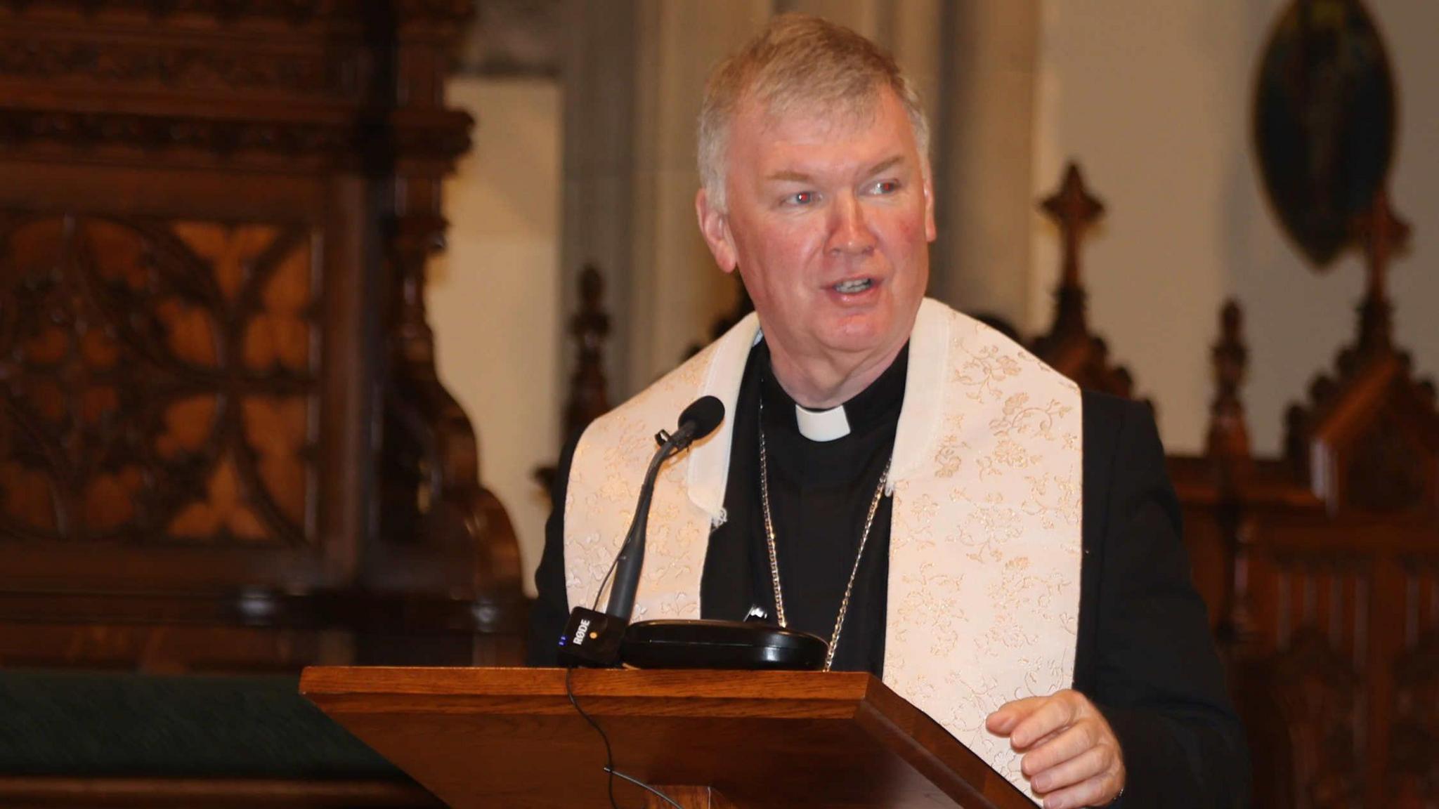Bishop Coll standing at a lectern. He has white hair and is wearing black priest attire with a white garment across his shoulders.