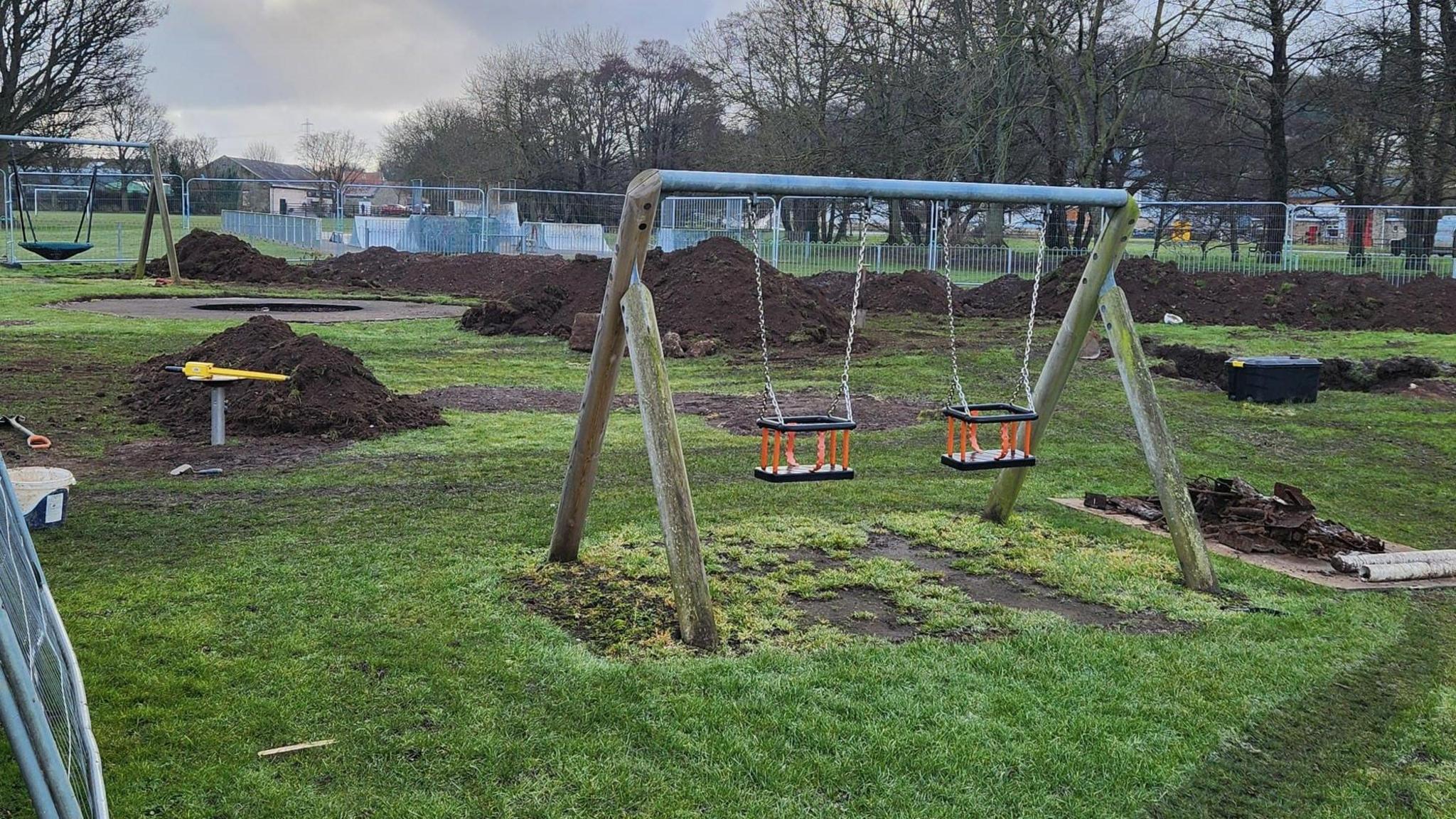 A set of swings is in the foreground of the picture and behind there are large mounds of dug-up earth. Fencing is all round the site.