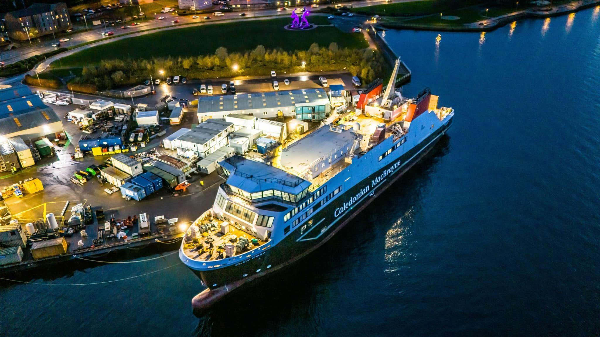 A large ship, lit up brightly at dusk, moored beside the Ferguson Marine shipyard with the large The Shipbuilders statue lit up in purple in the distance          