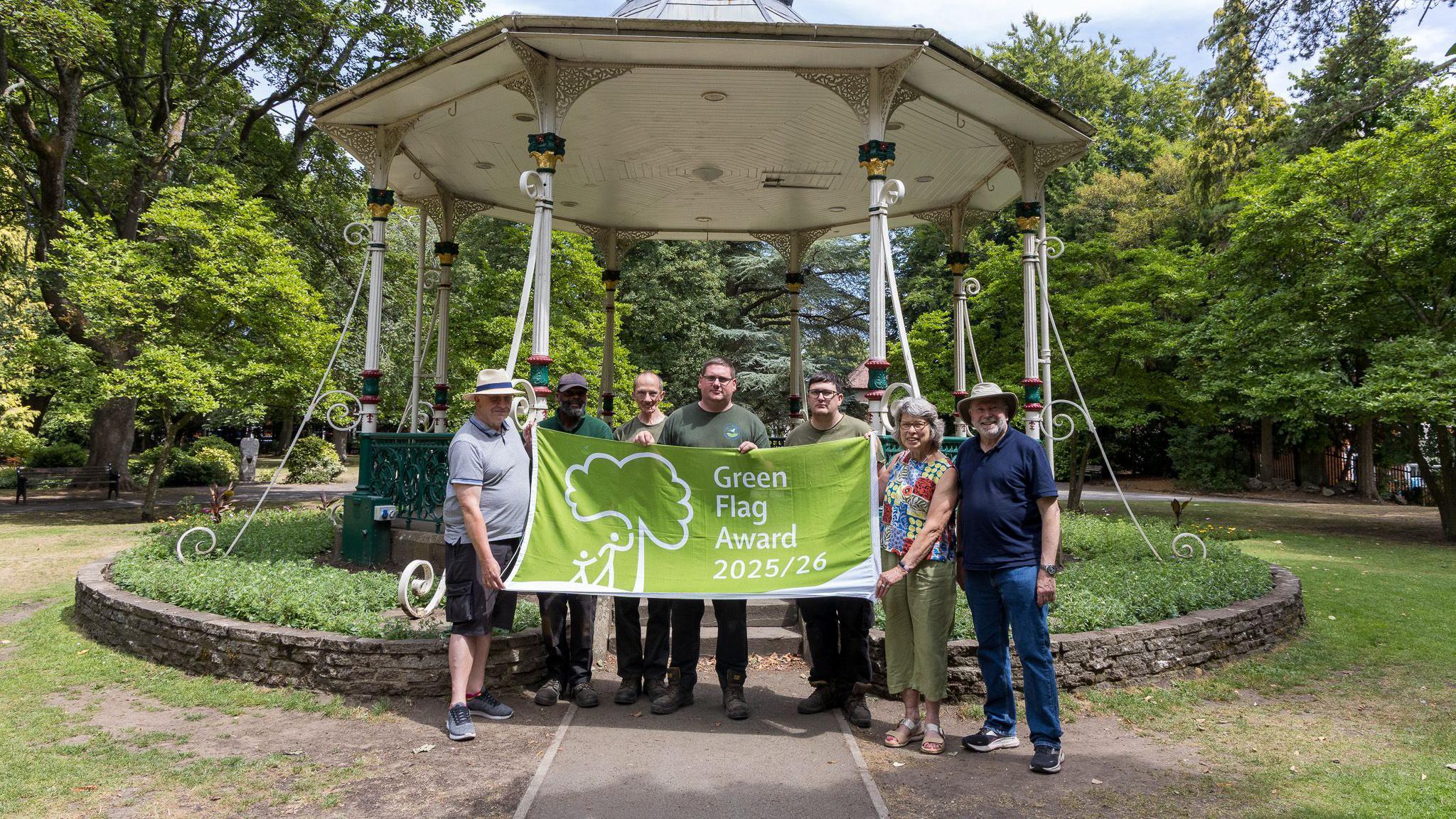 A group of people hold a green banner with the words "Green Flag Award 2025/26" in front of a bandstand in a park. Leafy green trees can be seen in the background but the grass around the bandstand is patchy and dry in places.