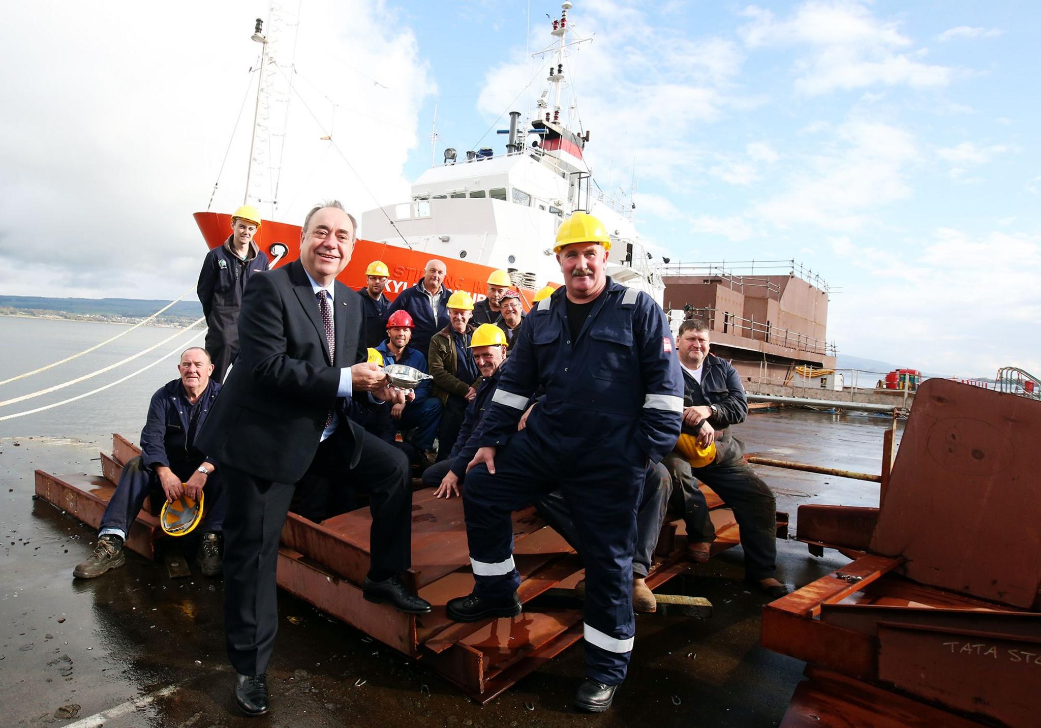 Alex Salmond, in a dark suit, stands next to union convener Alex Logan, in a boilersuit, with other Ferguson workers and a ship in the background