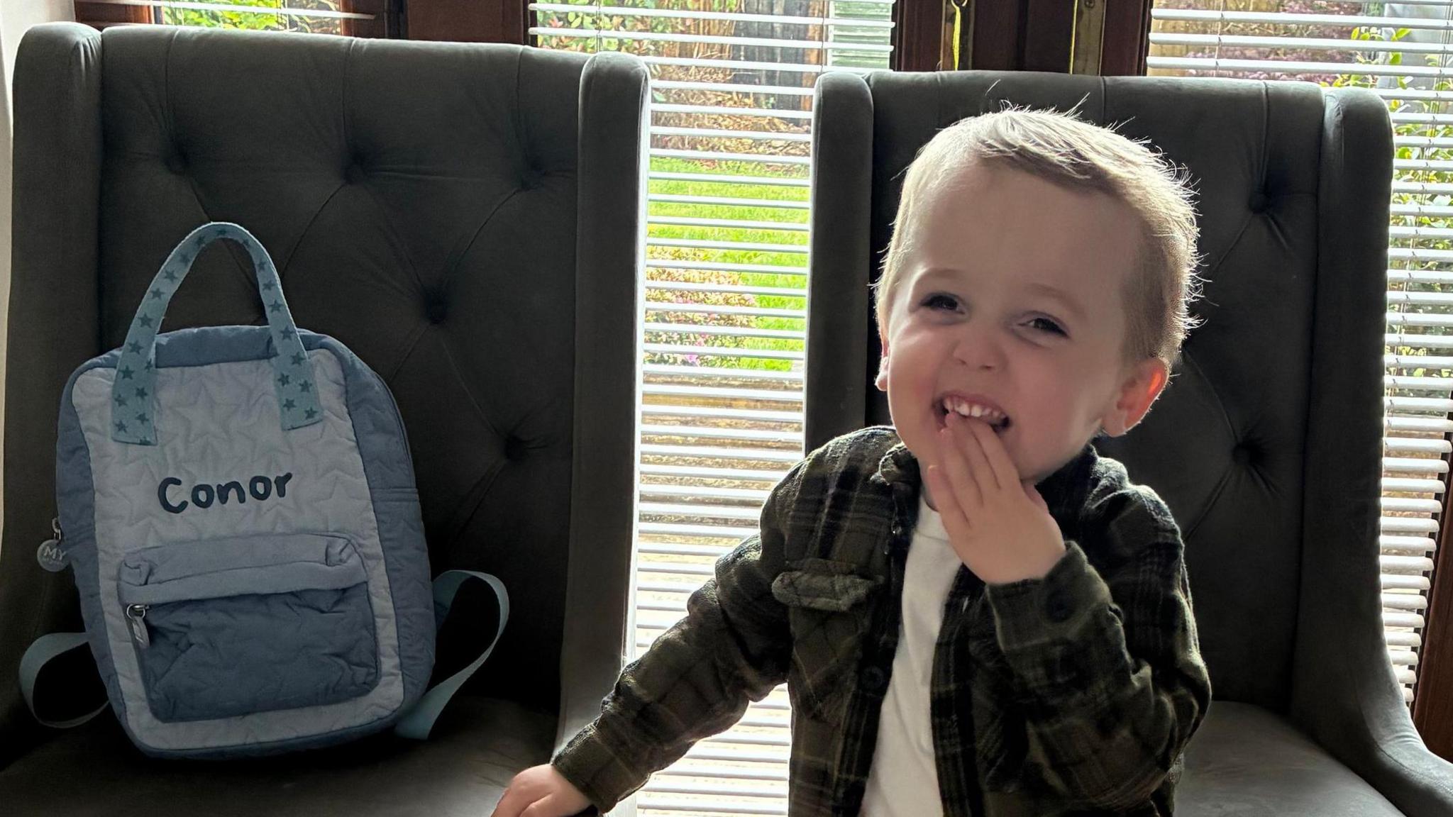 Conor grins and touch his mouth in front of a dark chair. A backpack with his name is on another dark chair next to him.