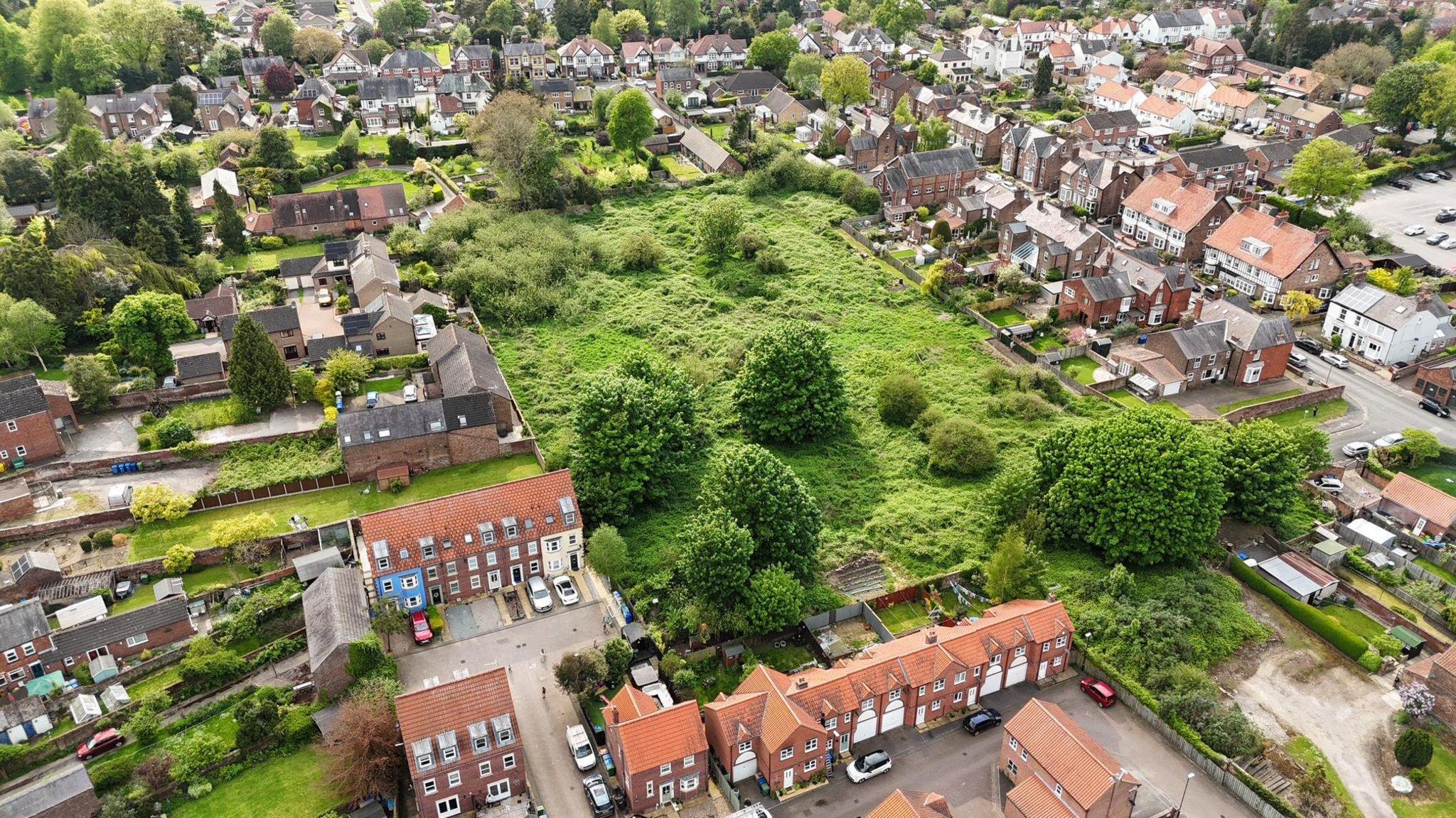 A bird's-eye drone shot of a green space in Driffield, which includes large bushes and trees. It is surrounded by residential houses and gardens. 