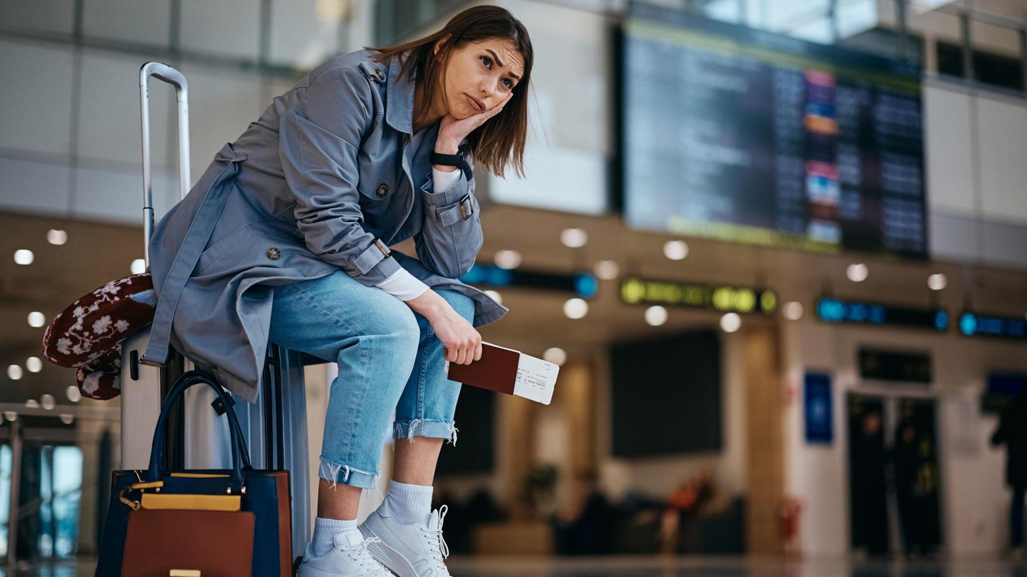 Young woman wearing blue jeans, a grey trenchcoat and white trainers sits on her suitcase at an airport, clutching her passport and ticket