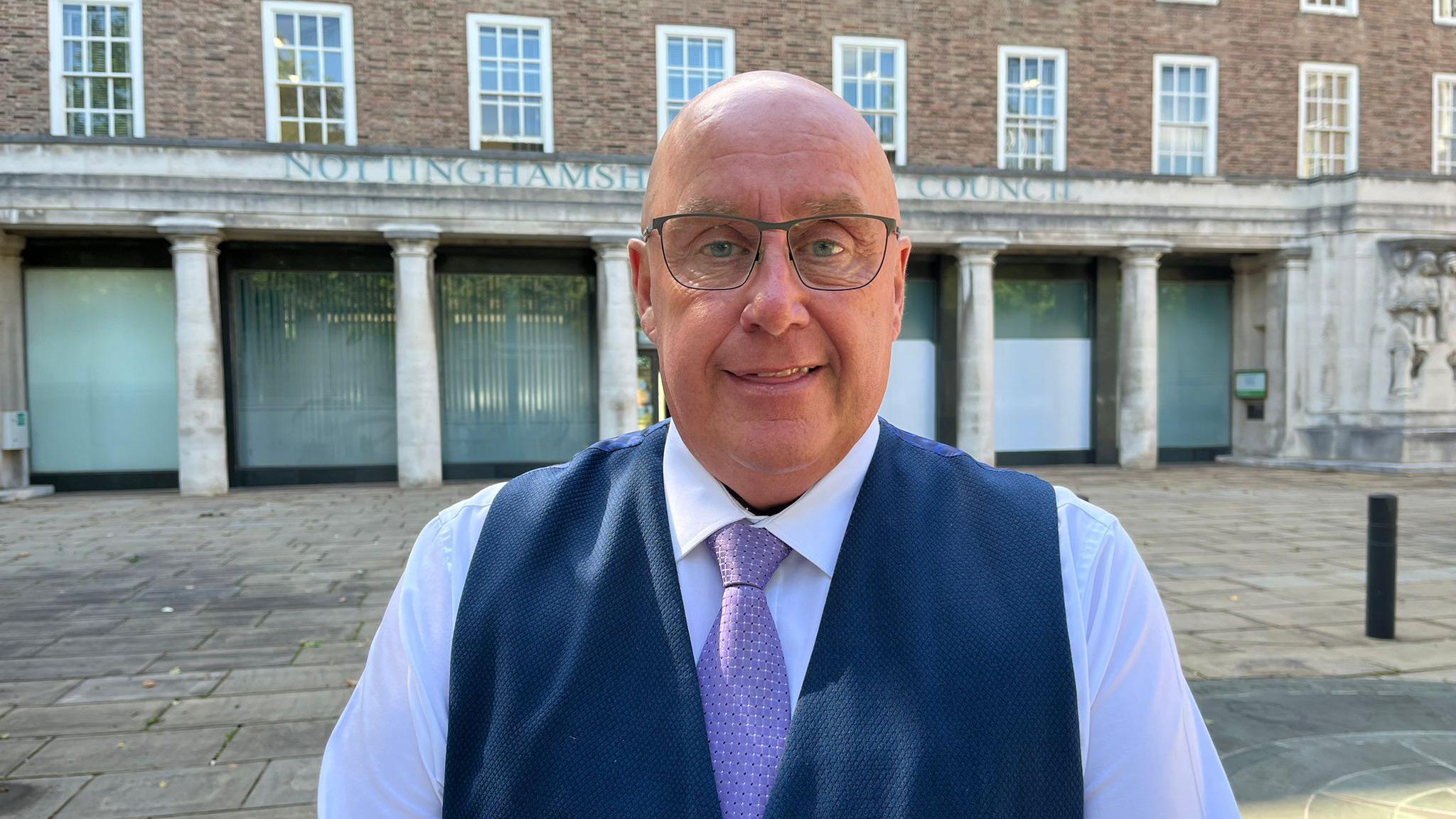 A man wearing glasses and a blue waistcoat and purple tie.