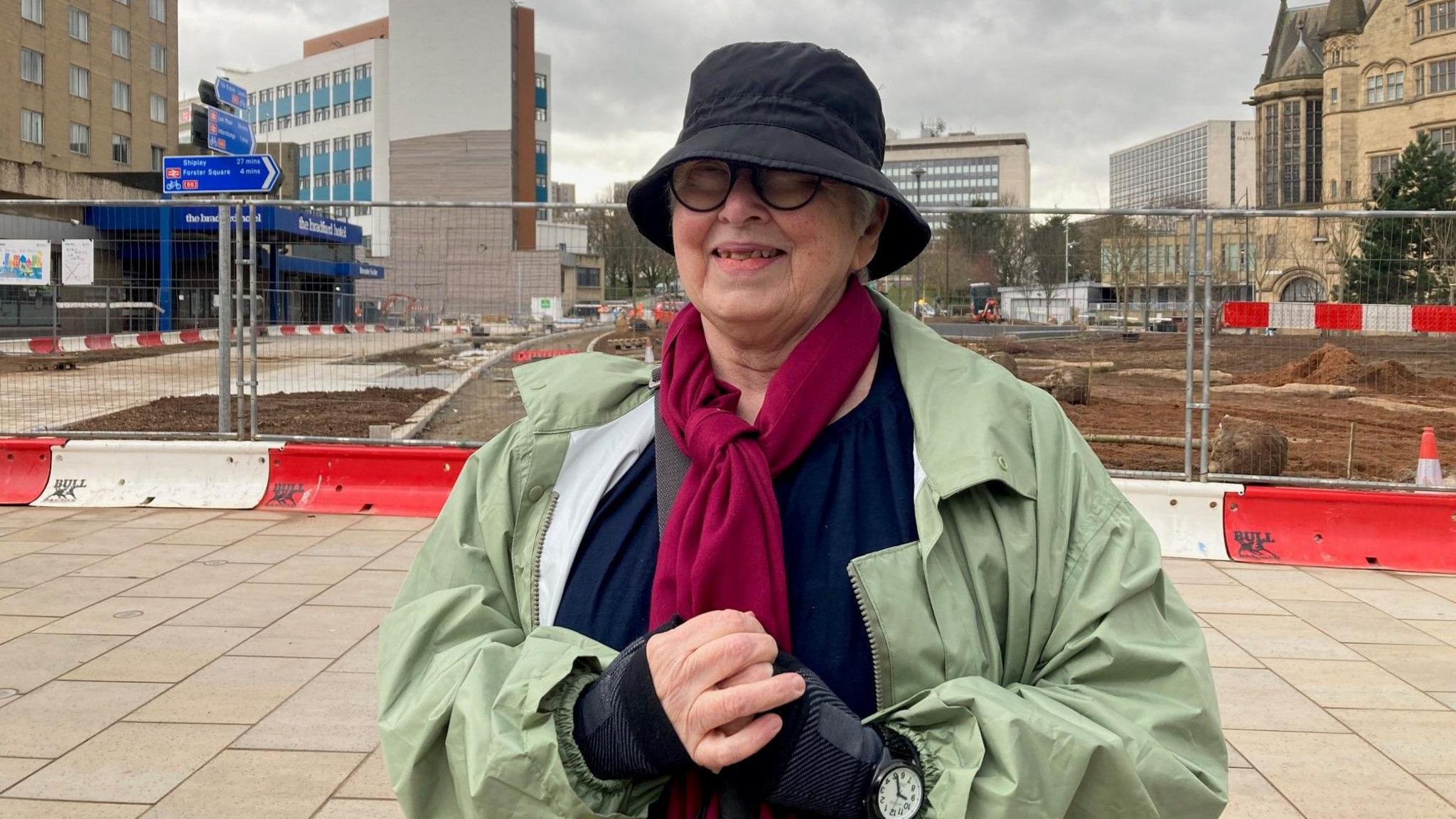 A woman wearing a hat, scarf and coat in front of a fenced-off building site with red and white safety barriers.