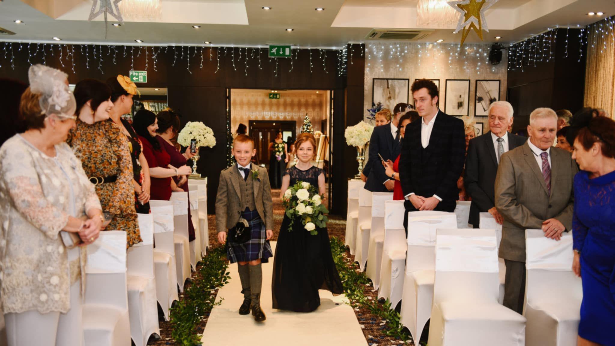 A young boy in a kilt and a girl holding flowers walk down the aisle at a wedding, watched by guests