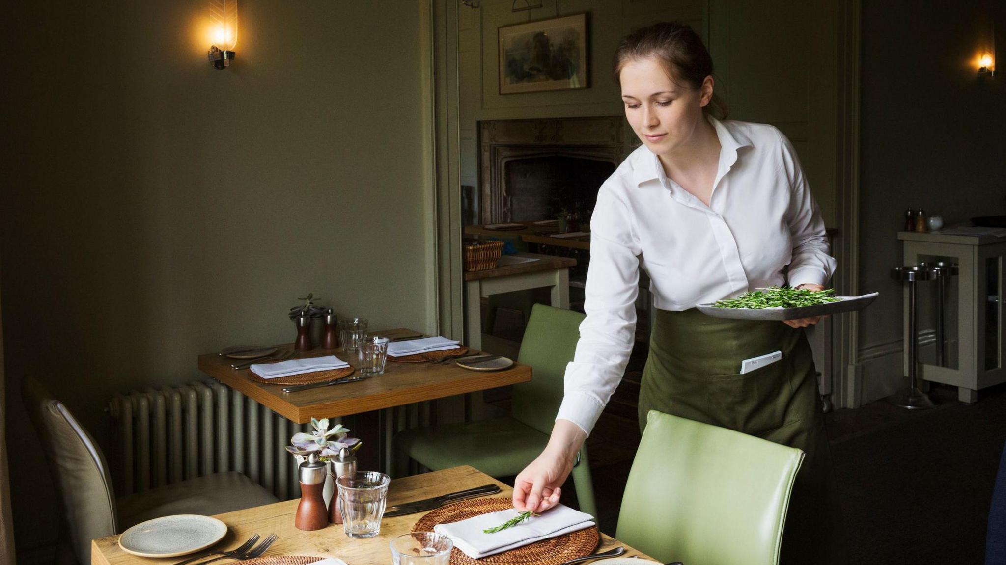 Female restaurant worker setting a table. She is wearing a white shirt and green apron carrying a plate. 