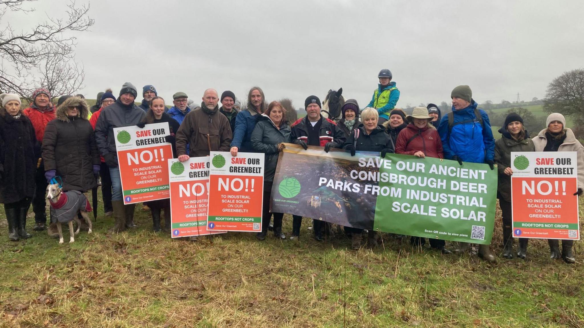 A group of people wearing winter coats and hats, huddle together in a field holding banners which say 'Save Our Greenbelt  NO!! to industrial scale solar on our greenbelt'