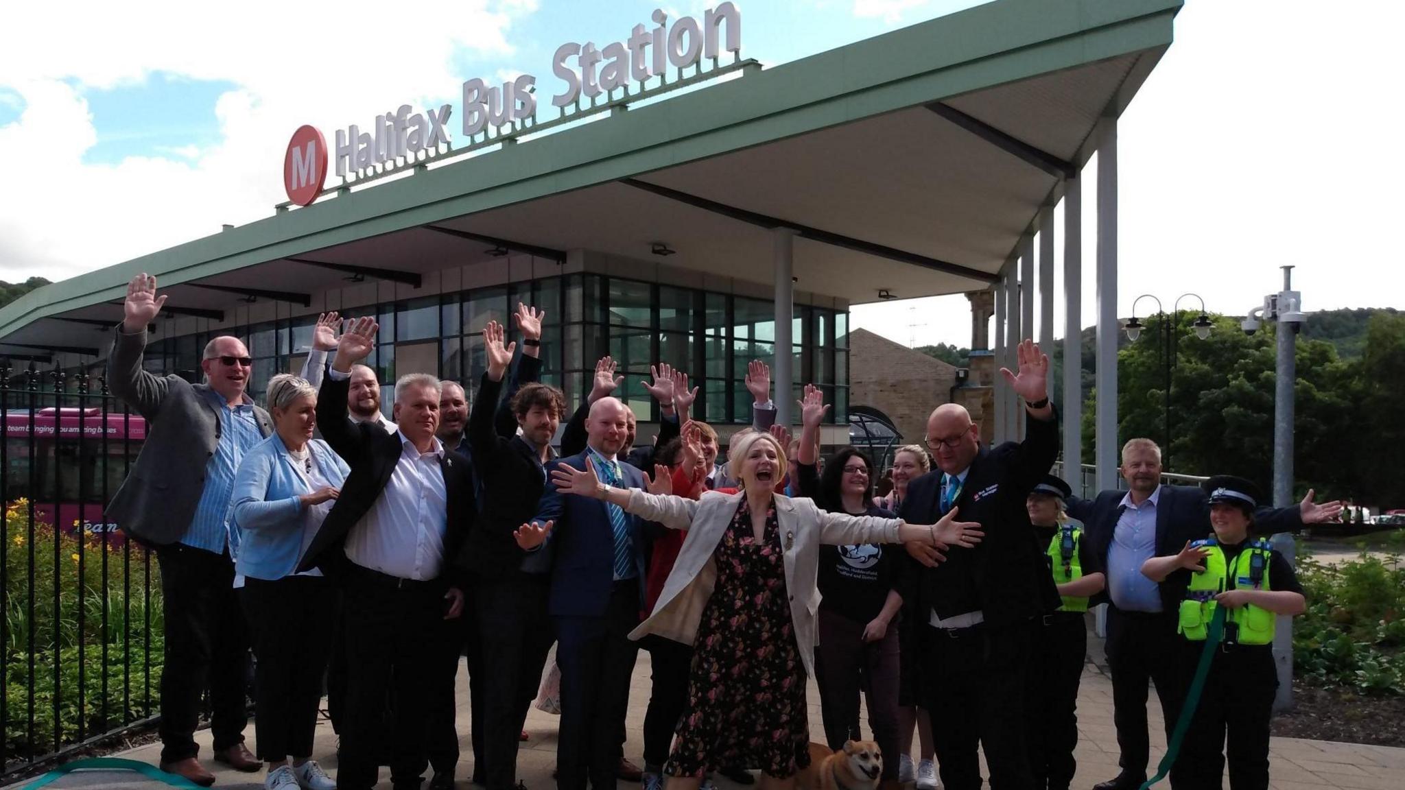 West Yorkshire Mayor Tracy Brabin officially opens the new bus station. A group of about 20 people stand in front of Halifax Bus Station - they are waving. Tracy Brabin is in the foreground with her arms wide. 