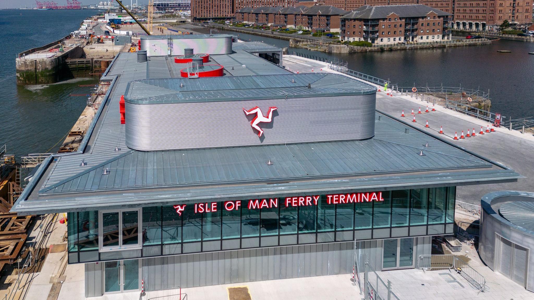 Aerial shot of the front of the ferry terminal, which has Isle of Man Ferry Terminal written across the windows on the second floor and a Manx triskelion on the roof above. There is water either side and red brick building to the right behind it.