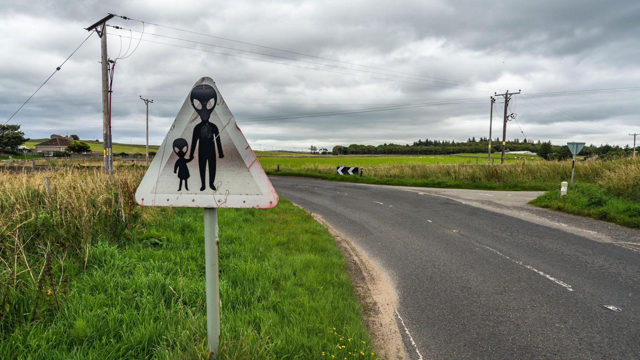 A road sign showing two aliens in silhouette crossing a road. They have large heads with huge almond-shaped eyes. The larger - presumably adult - alien is holding the hand of a small, presumably infant, alien. The sign is weathered, triangular, and by the side of a bending road with a small bungalow in the background.