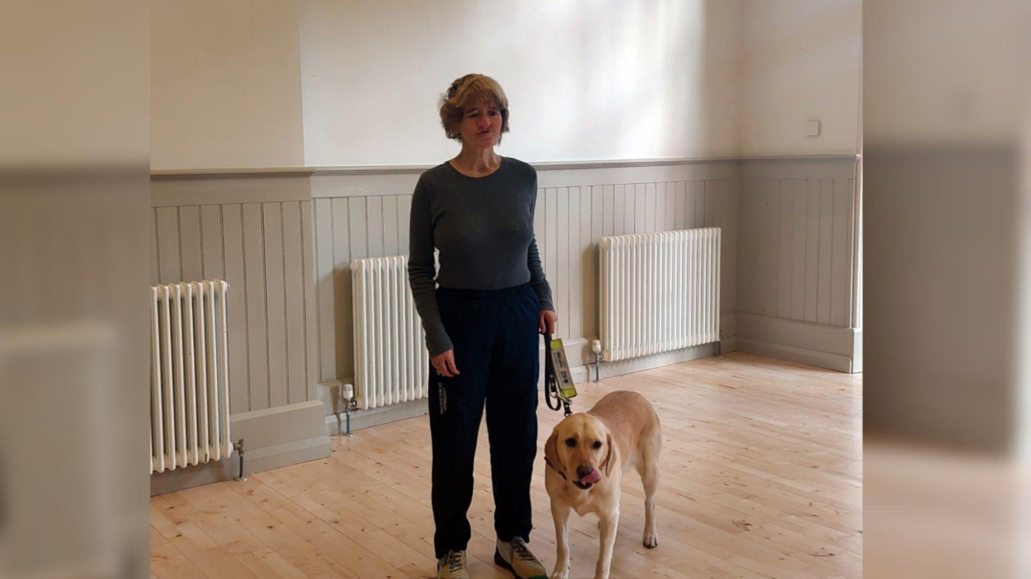 Angela Hope is standing in the dance studio on a light wooden floor. She has short blonde hair and is wearing a long-sleeve grey top and dark trousers. She is holding the harness of a yellow Labrador guide dog. Behind her is a white wall with pale wooden paneling and three white radiators along the wall.